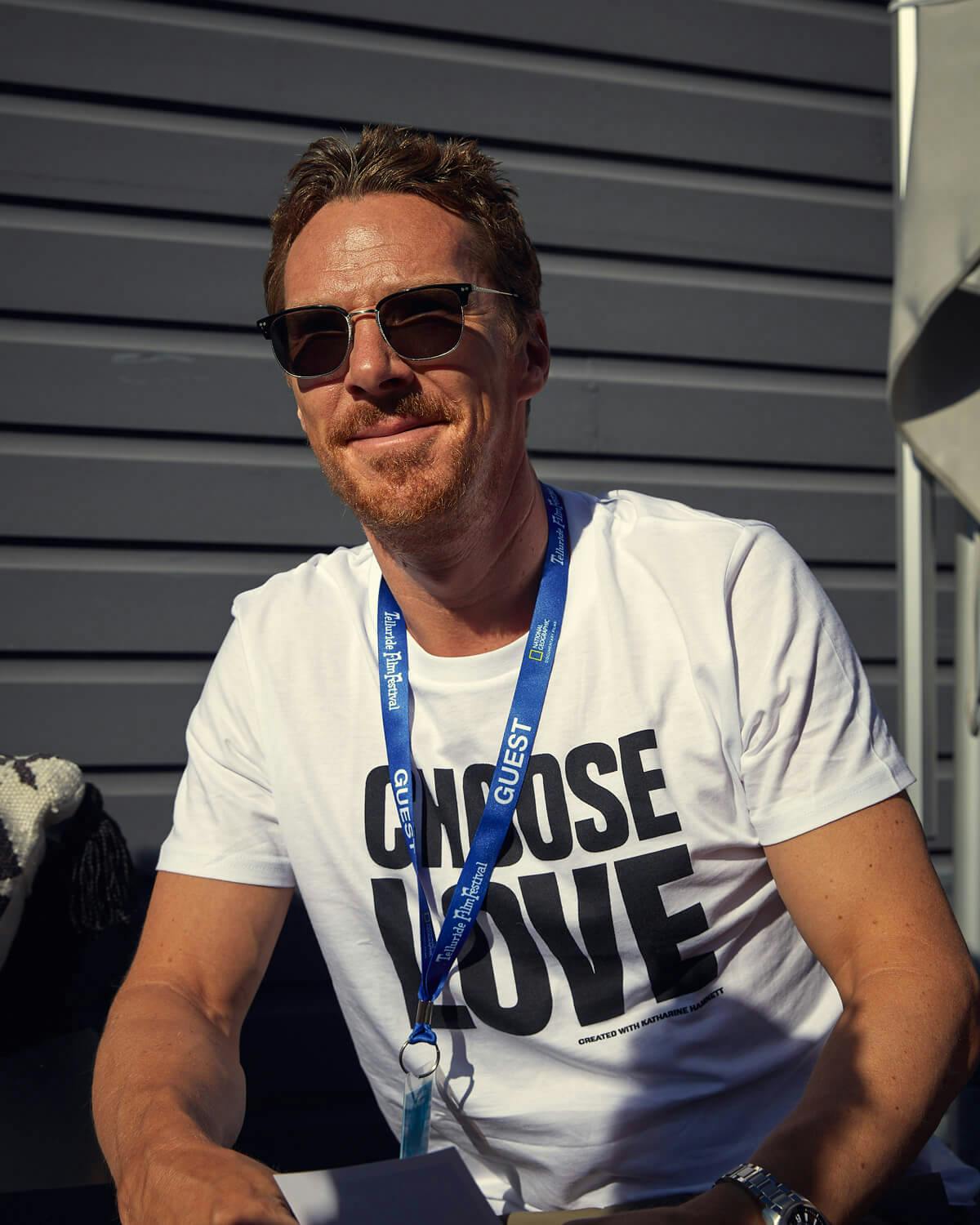 Benedict Cumberbatch wears a white t-shirt reading “Choose Love” and wearing sunglasses sits outside at the Telluride Film Festival, 2021.
