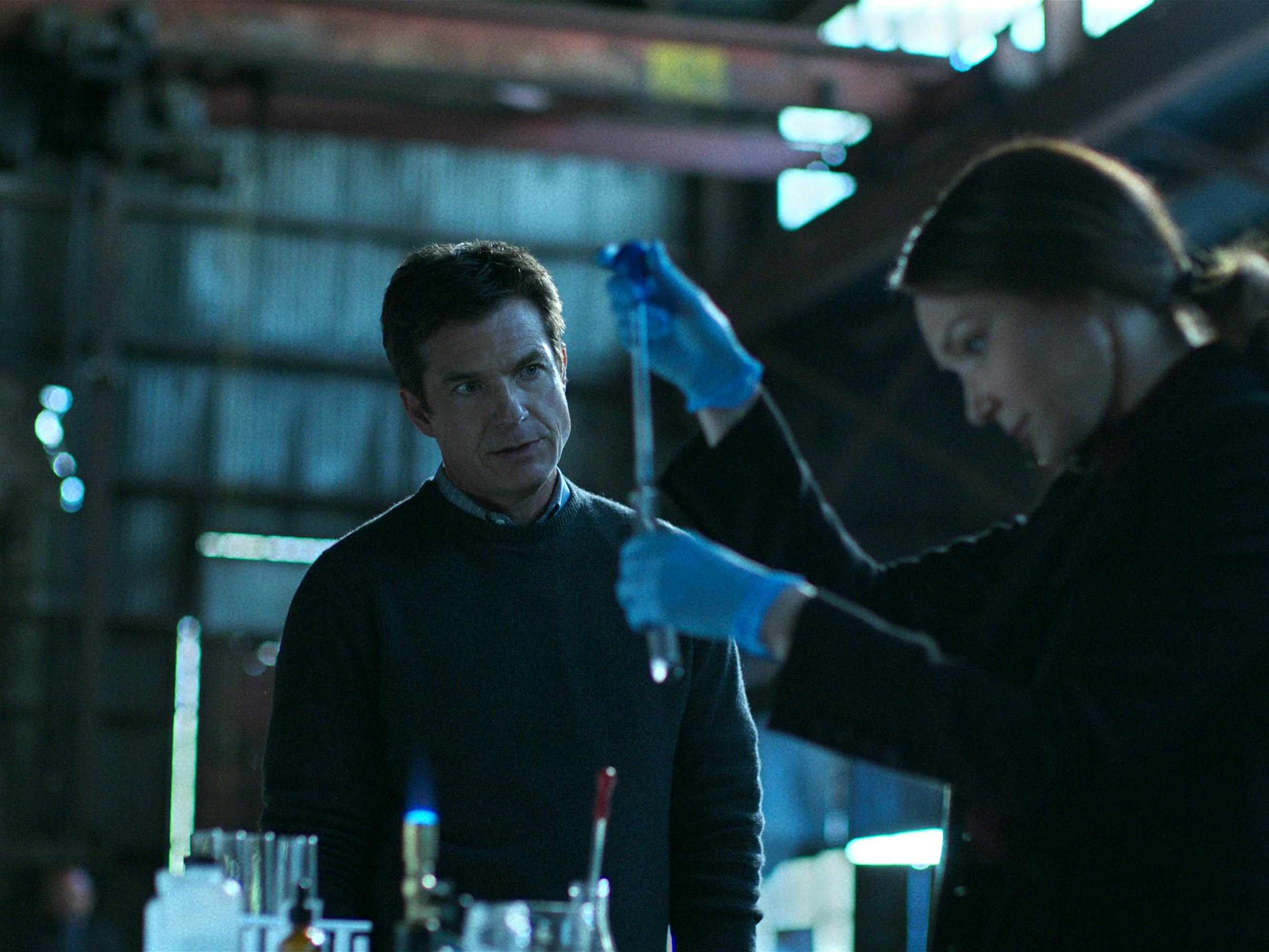 Jason Bateman and Katrina Lenk stand around vials and a small open flame. Bateman and Lenk both wear dark sweaters.