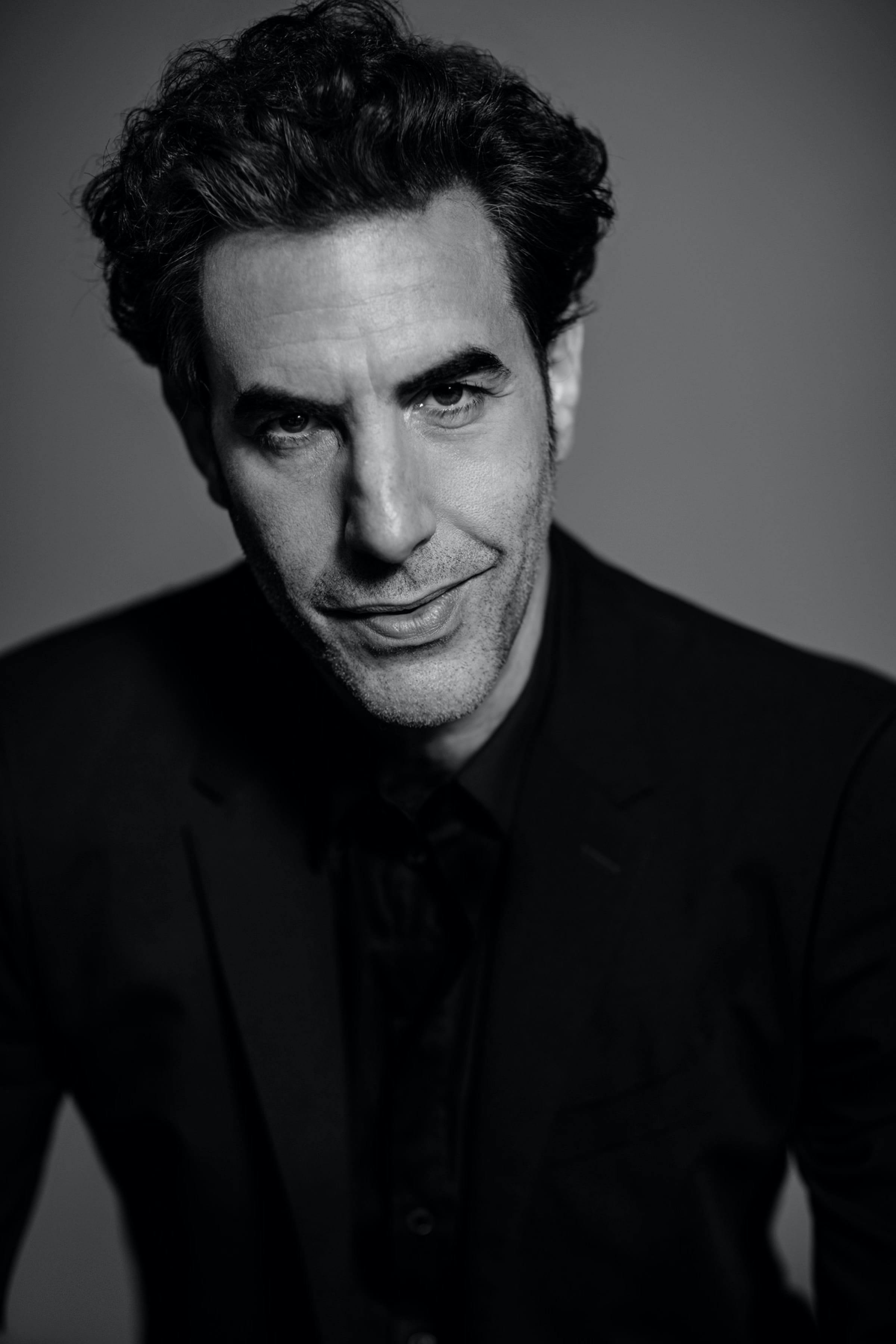 A black-and-white shot of Sacha Baron Cohen in a black suit smiling slightly looking at the camera.