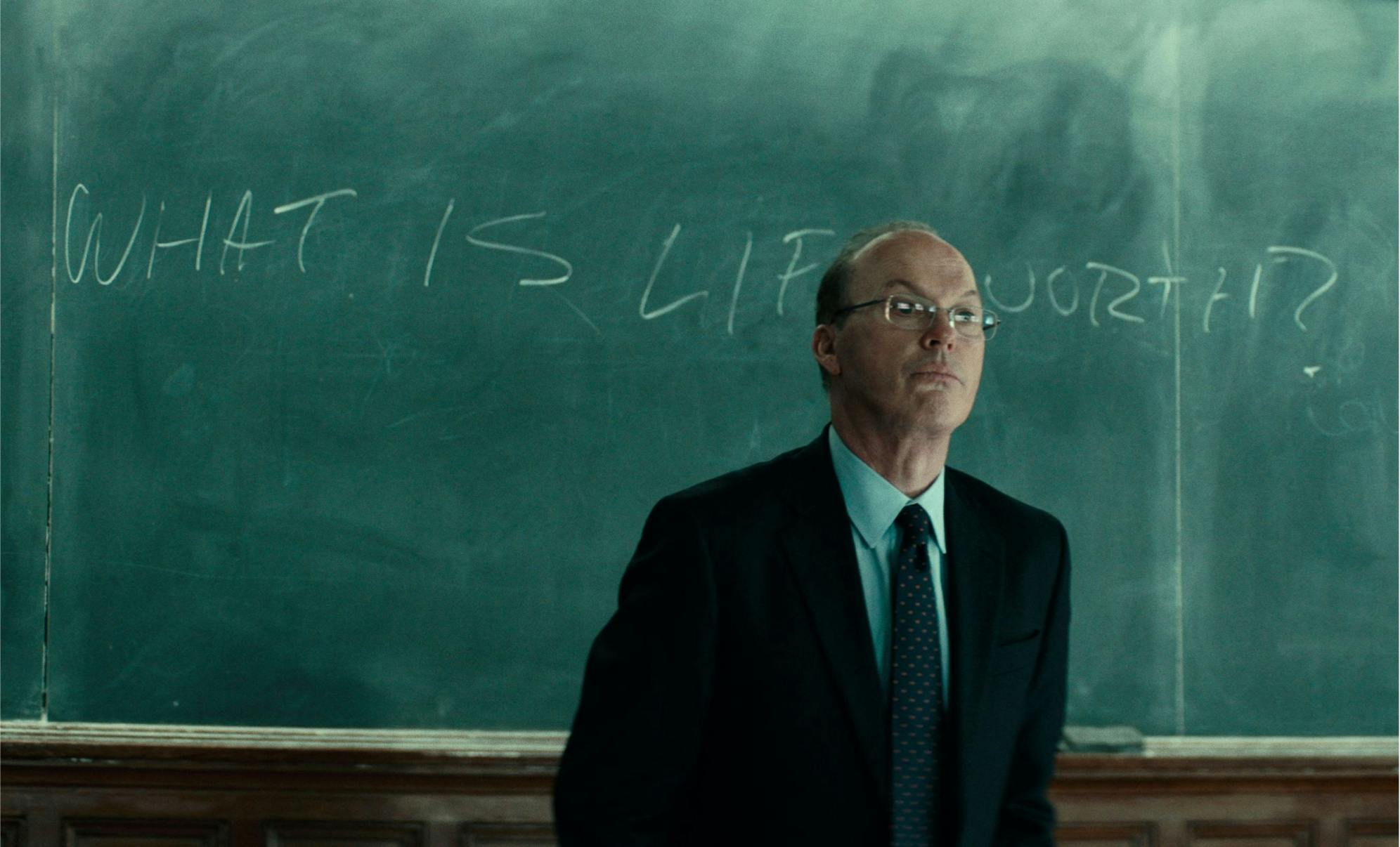Kenneth Feinberg (Michael Keaton) stands in front of a chalkboard with the words: “What is life worth?” He wears a suit and glasses.
