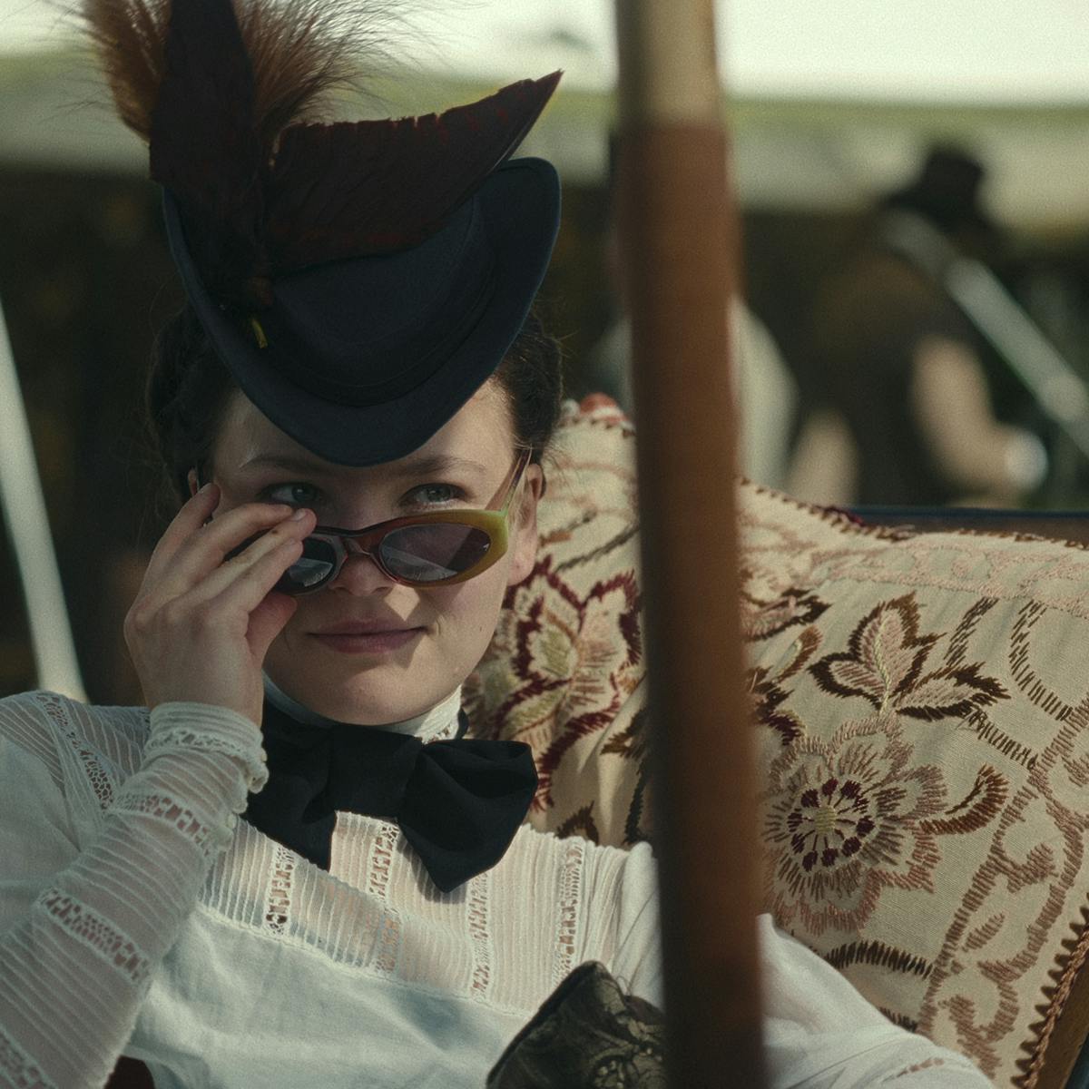 Elisabeth (Devrim Lingnau) wears a white shirt and black necktie, as well as a feathered black cap and sunglasses.