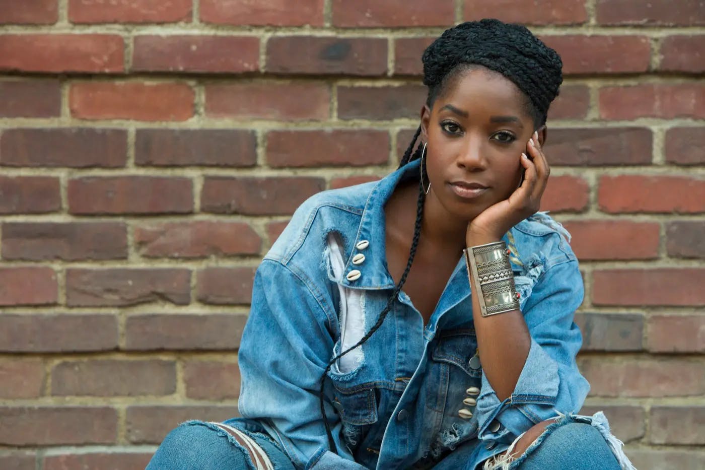 Joelle Brooks (Ashley Blaine Featherson) sits casually in front of a brick wall, her elbow resting on her knee and cupping her face.  She wears ripped jeans and a denim jacket decorated with shells on the collar and pocket.  A chunky bronze cuff bracelet adorns her wrist, complementing her large hoop earrings.