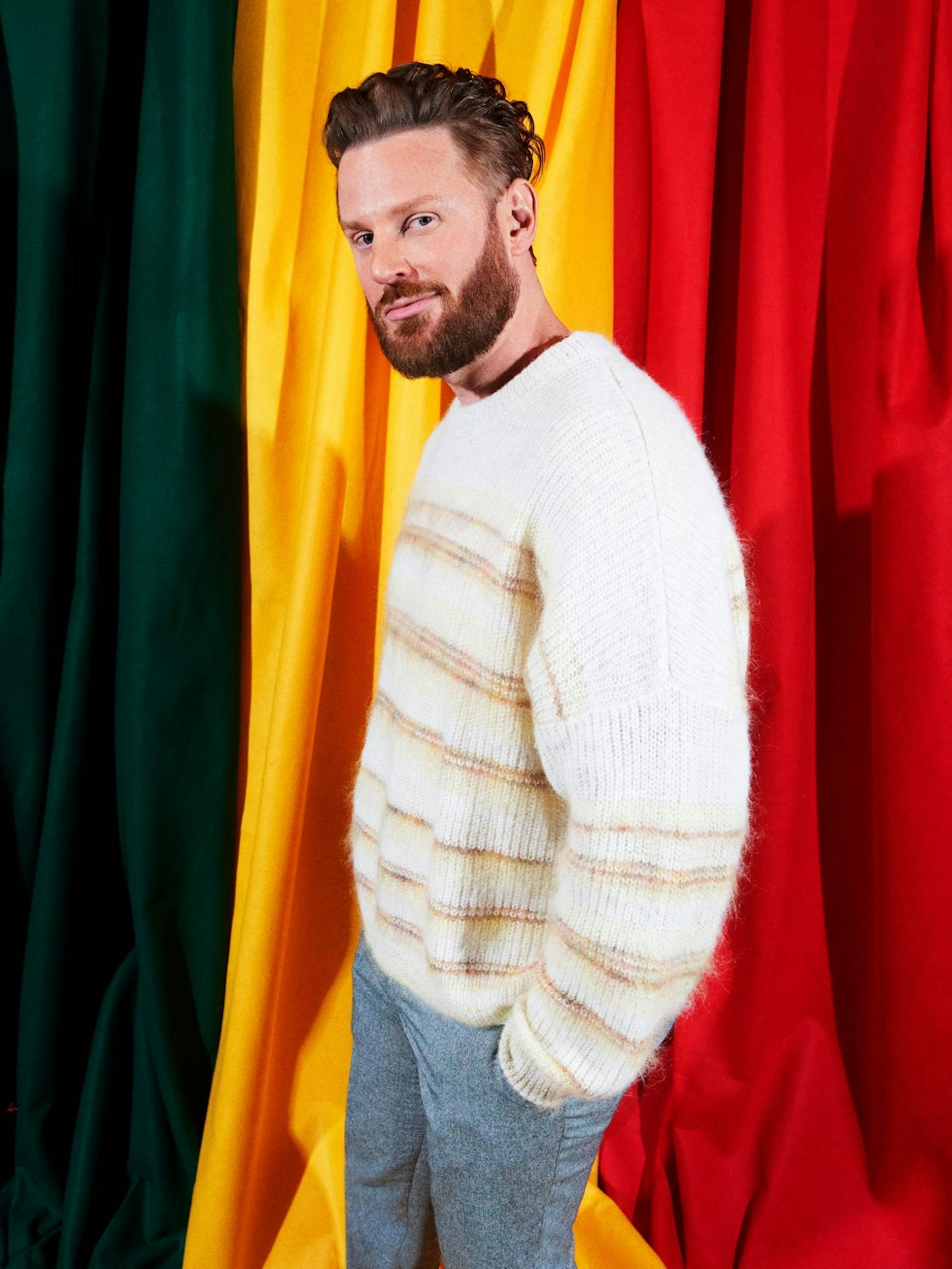 Bobby Berk stands against a green, yellow, and red clothed backdrop. He wears a furry white and orange sweater, which matches his well trimmed beard and slicked back hair.