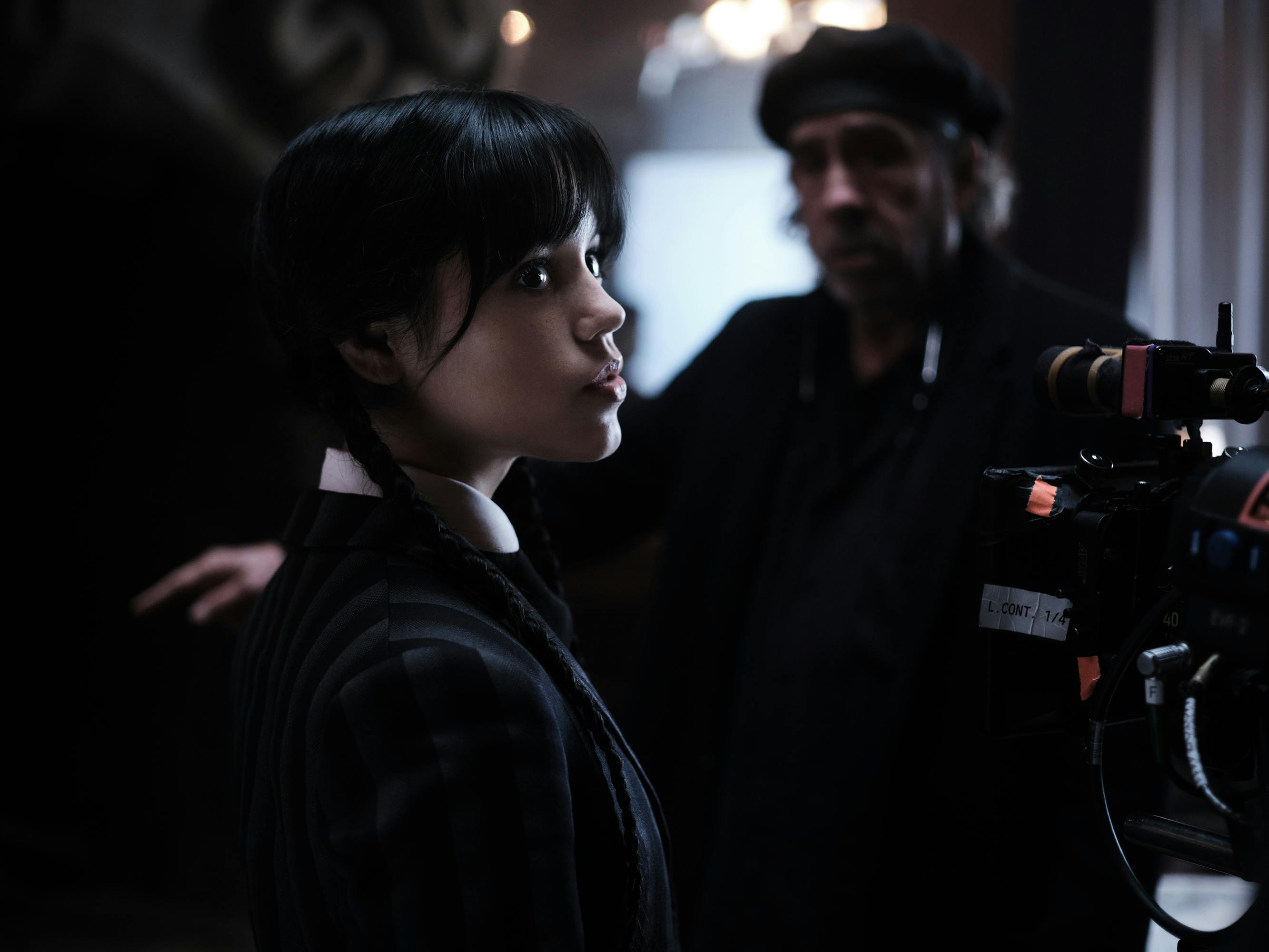 Jenna Ortega looks at something off-camera. Tim Burton is out-of-focus in the background. 