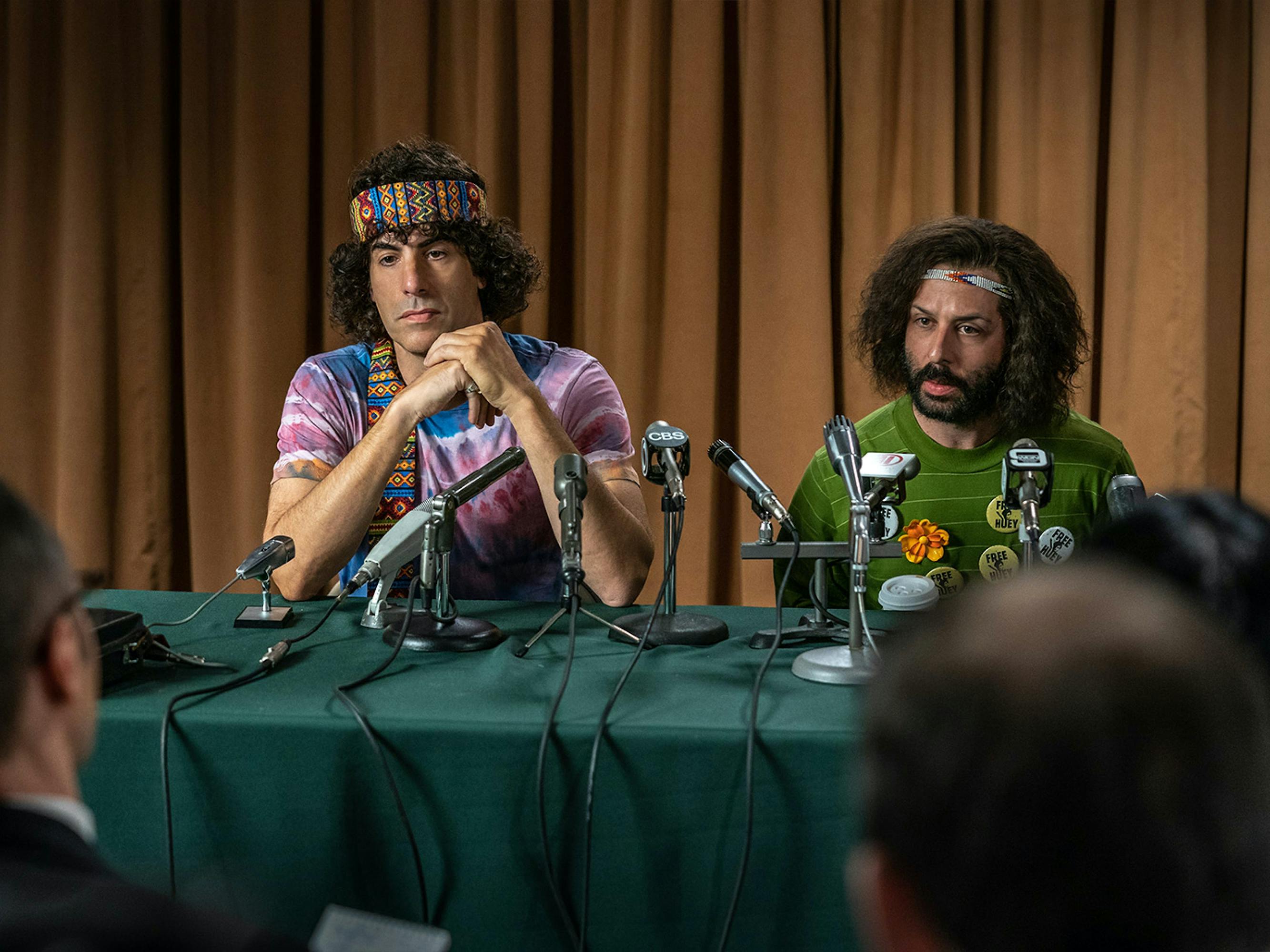 Cohen and Strong sit behind a table of microphones facing reporters. Cohen wears a colorful headband and a tie-dyed shirt. Strong’s headband is thinner, and his shirt is covered in Black Panther Party pins.