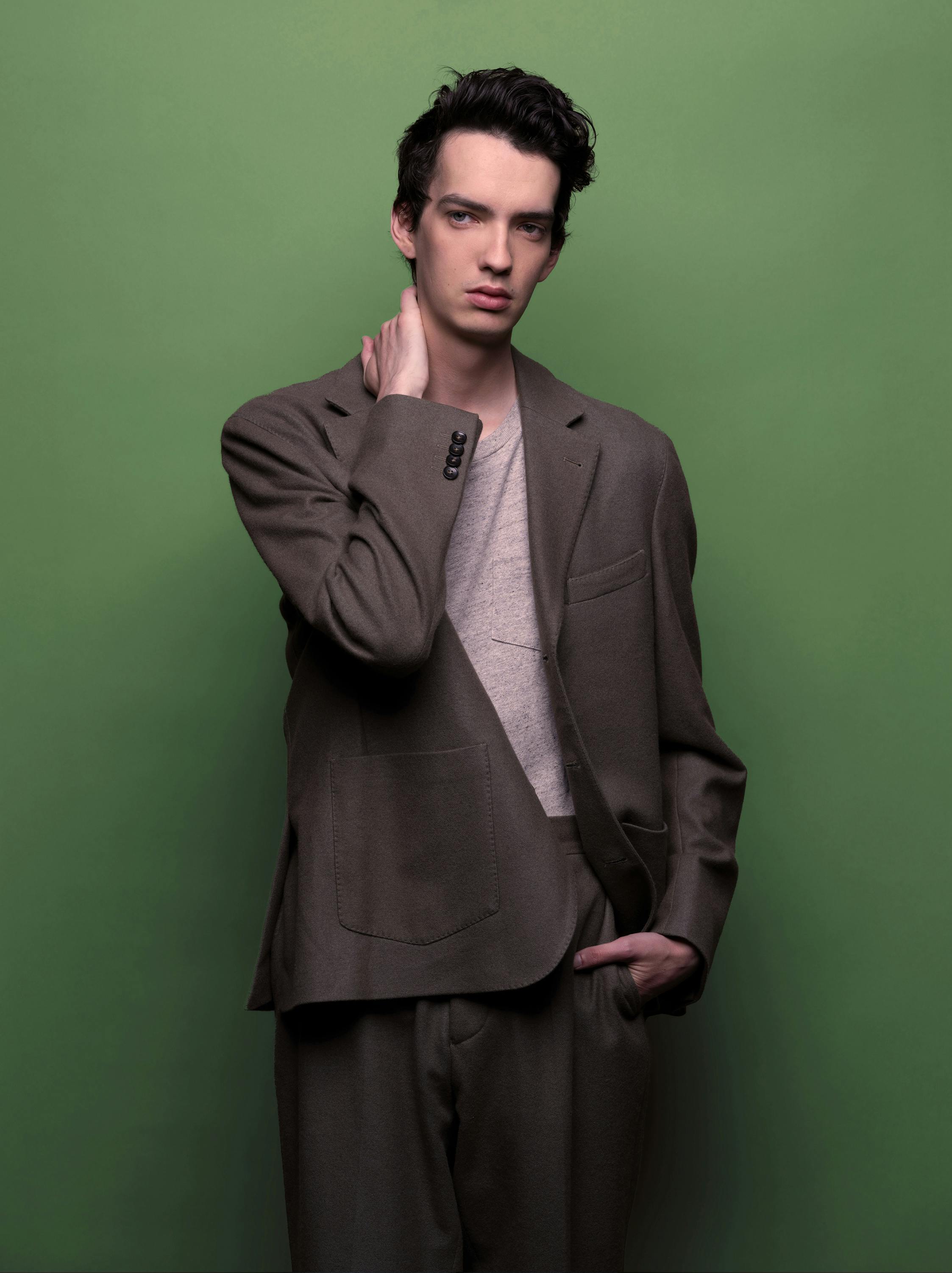 Kodi Smit-McPhee wears a grey brown suit and stands against an olive green wall. He holds one hand on his neck and looks at something off screen.