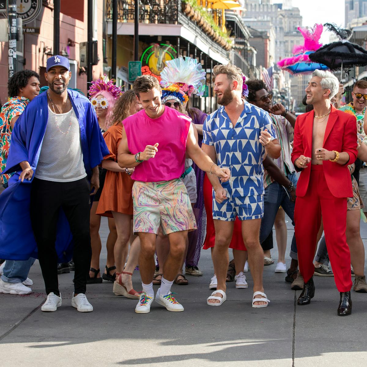 The Fab Five stand in front of a crowd of New Orleans heroes. Karamo wears black pants, a white tank top, a blue button-down, and a blue hat. Antoni wears a pink shirt and patterned shorts. Bobby wears a blue and white patterned romper. Tan wears a red suit and dark shoes. Jonathan wears a colorful strapless dress and heels. 