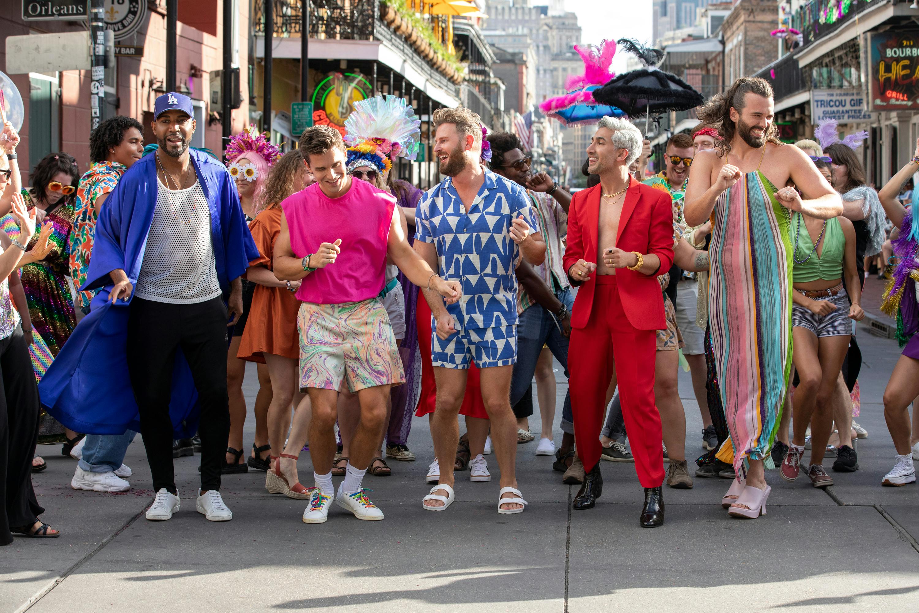 The Fab Five stand in front of a crowd of New Orleans heroes. Karamo wears black pants, a white tank top, a blue button-down, and a blue hat. Antoni wears a pink shirt and patterned shorts. Bobby wears a blue and white patterned romper. Tan wears a red suit and dark shoes. Jonathan wears a colorful strapless dress and heels. 