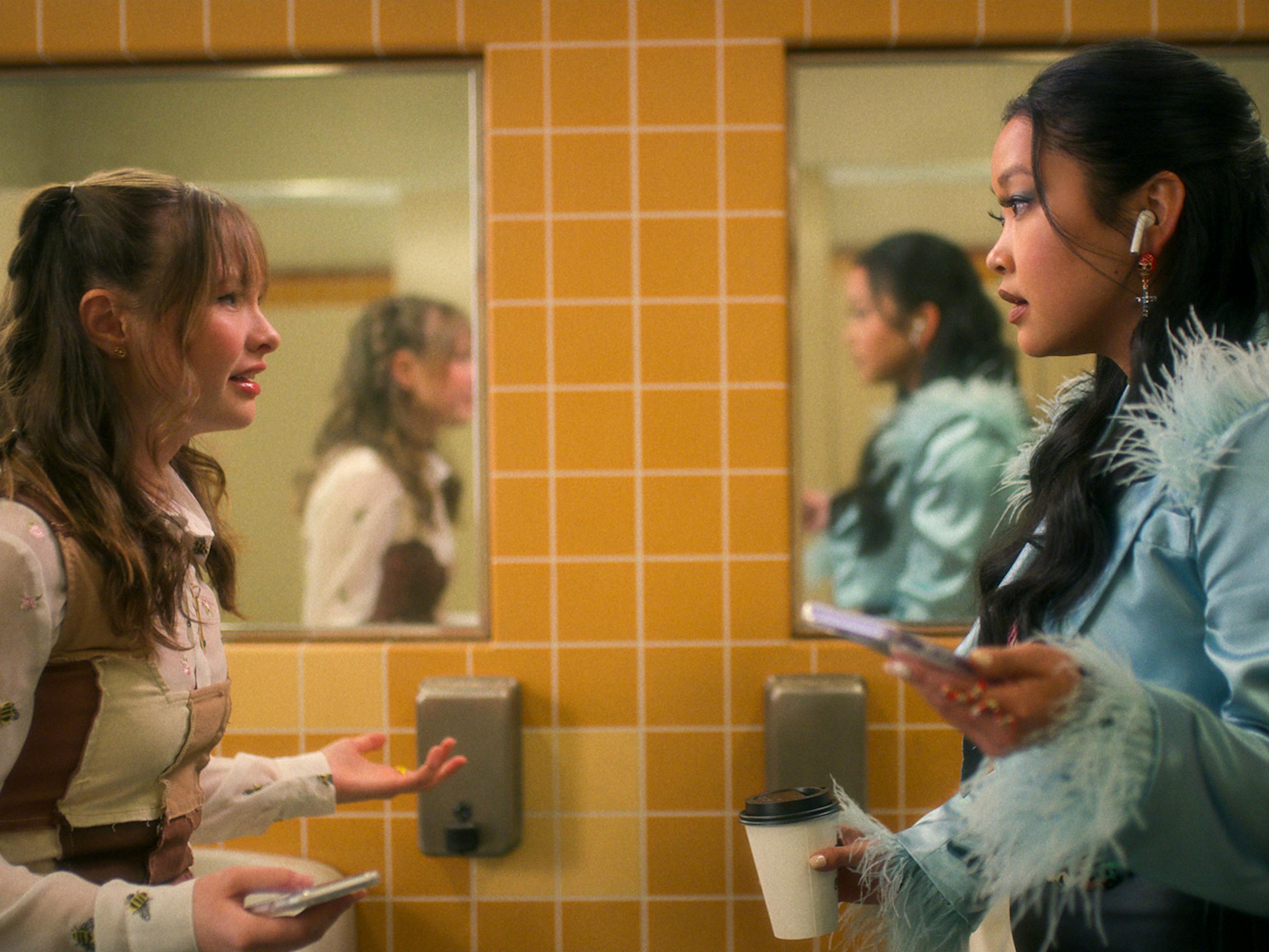 Gia (Zoe Margaret Colletti) and Erika (Lana Condor) wear fabulous outfits in a yellow-tiled bathroom.