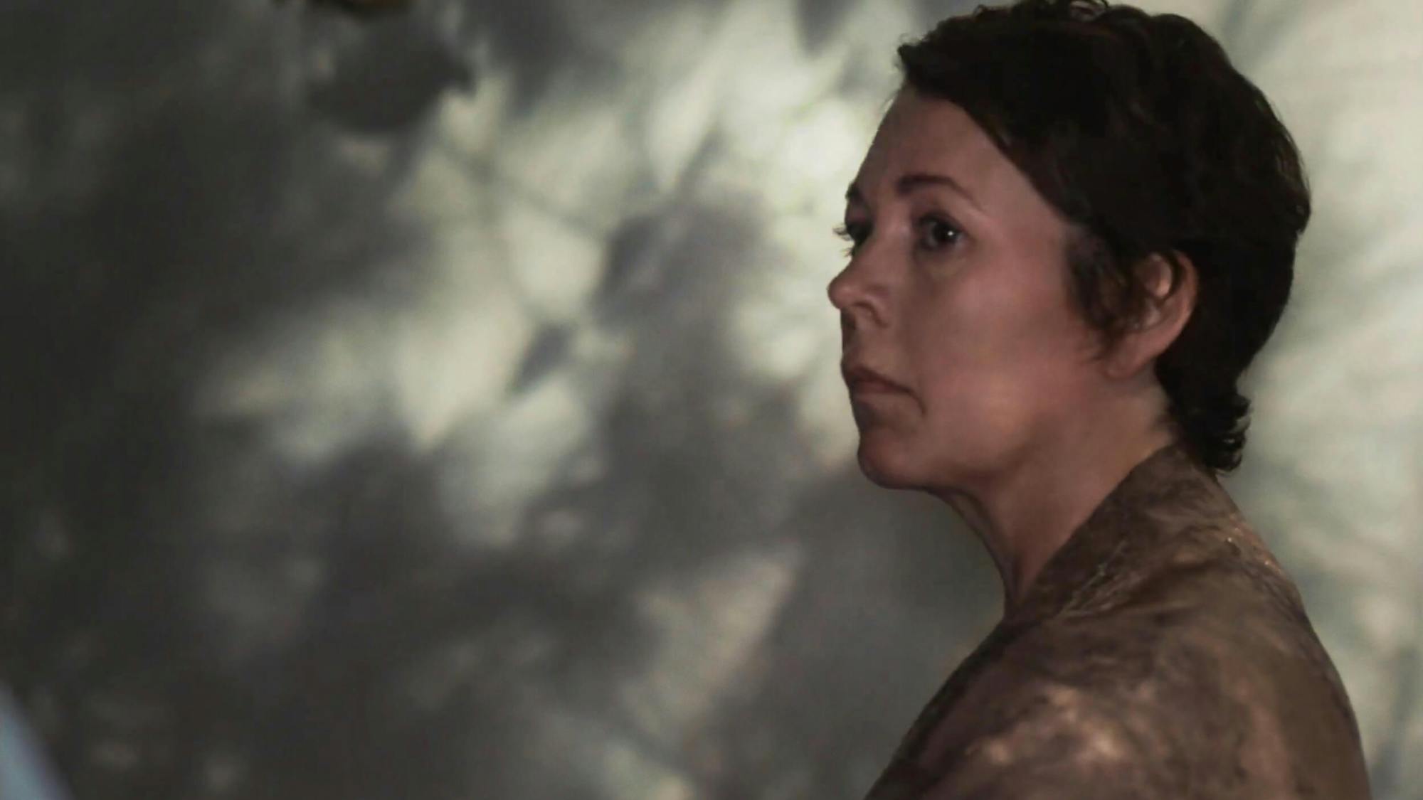Leda (Olivia Colman) wears a tan shirt. The wall behind her is shadowed with trees and leaves.