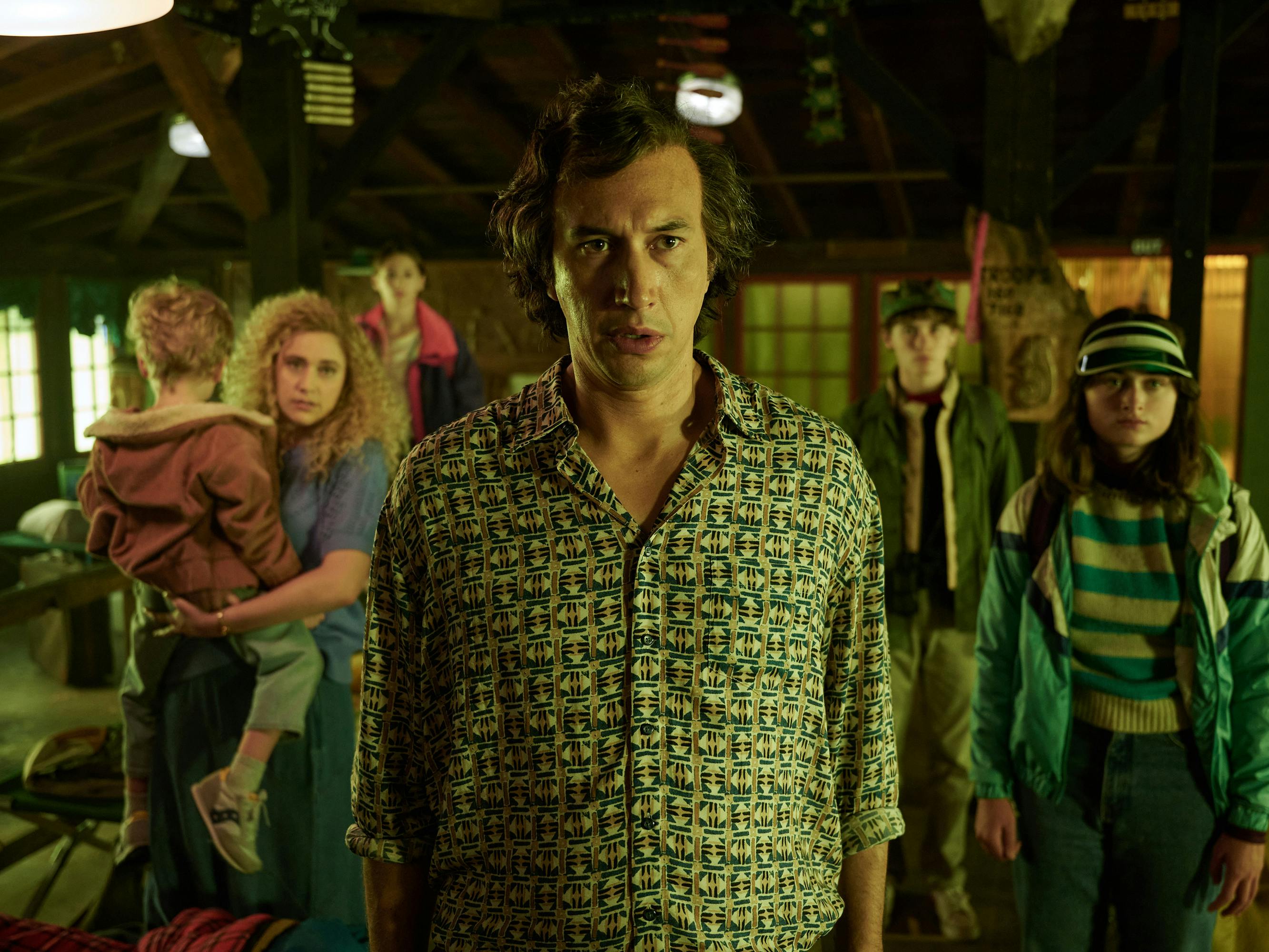 The Gladney family stand facing something offscreen. Greta Gerwig has a big perm and holds a baby, and Adam Driver faces the camera head on in a patterned shirt.