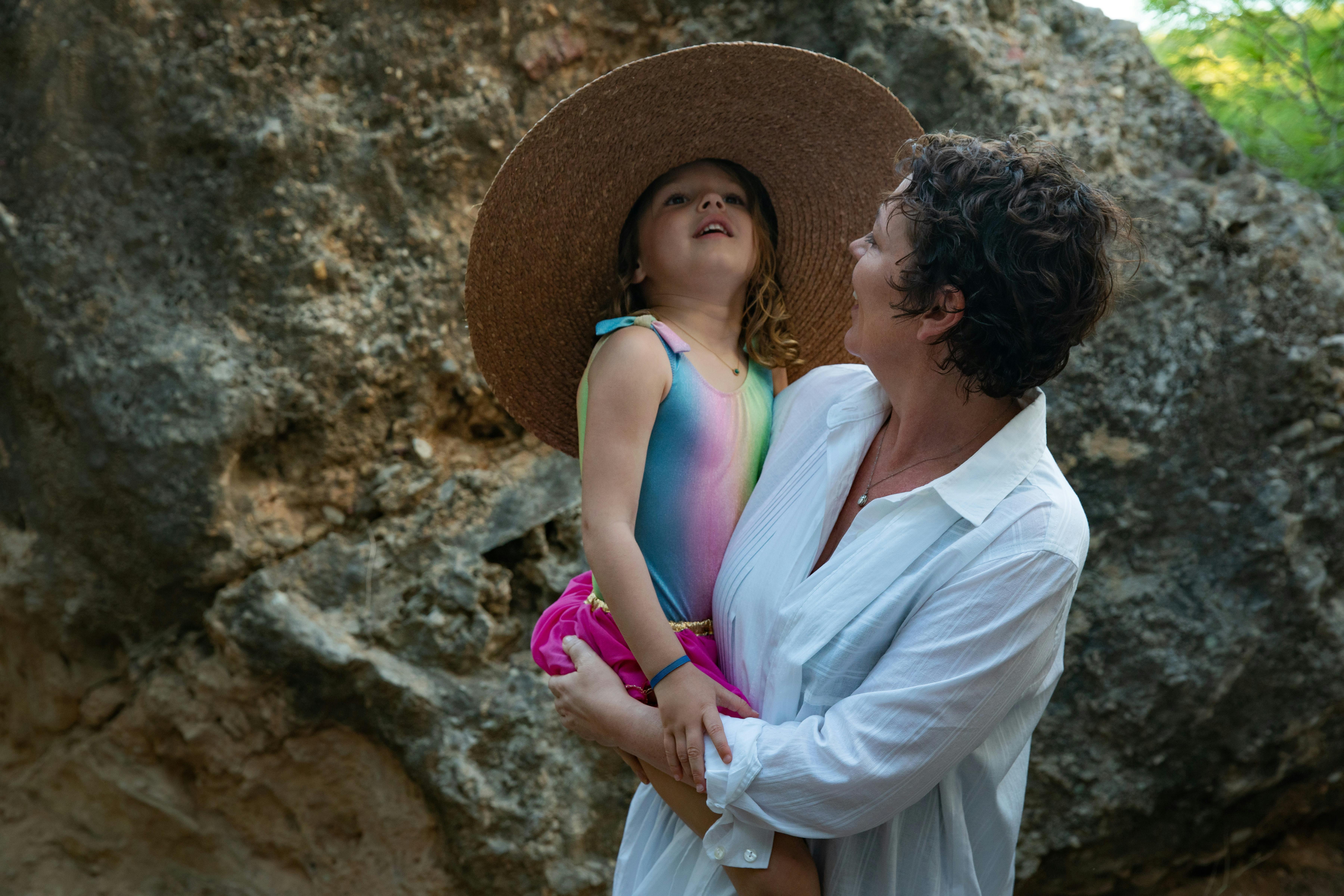 Olivia Colman holds Athena Martin by a big rock. Colman wears a big white buttoned down, Athena wears a colorful bathingsuit and a big straw hat.