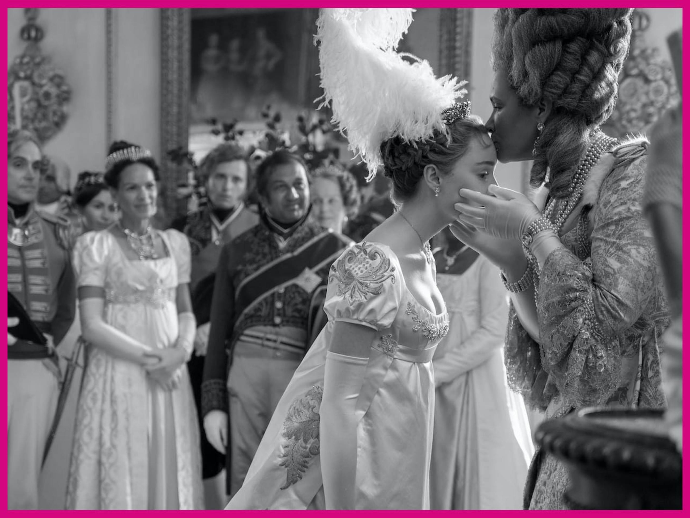 Daphne Bridgerton (Phoebe Dynevor) and Queen Charlotte (Golda Rosheuvel) embrace amidst a group of people. Queen Charlotte kisses Daphne’s forehead. The women are decked in shining dresses and gloves, and Daphne wears an impressive white plume atop her head.