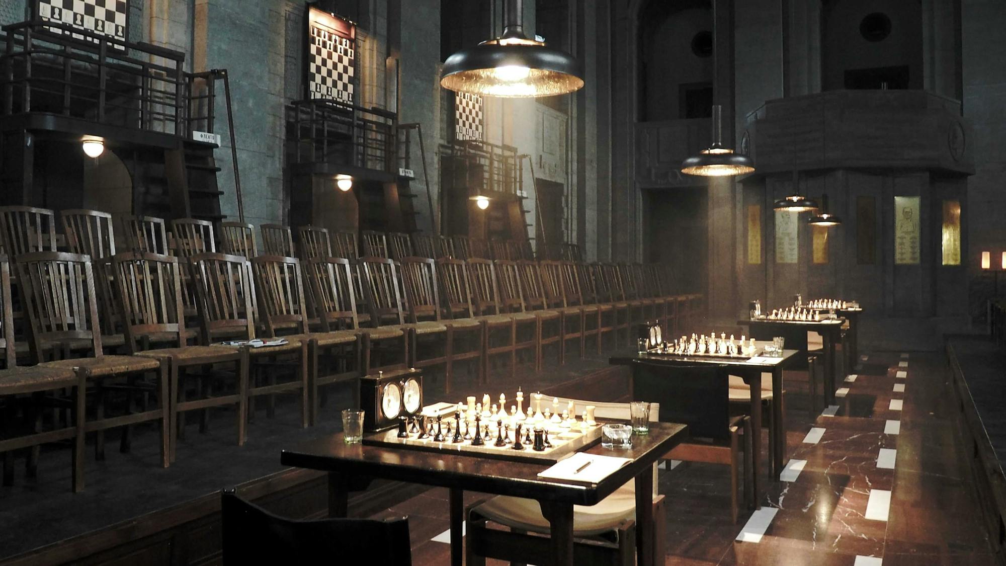 The Moscow chess tournament shot at The “Bear Chamber” in the Bärensaal contains rows of chairs, tables with chess boards and pieces, and shiny wooden floors. The dark scene is lit with overhead chandeliers.