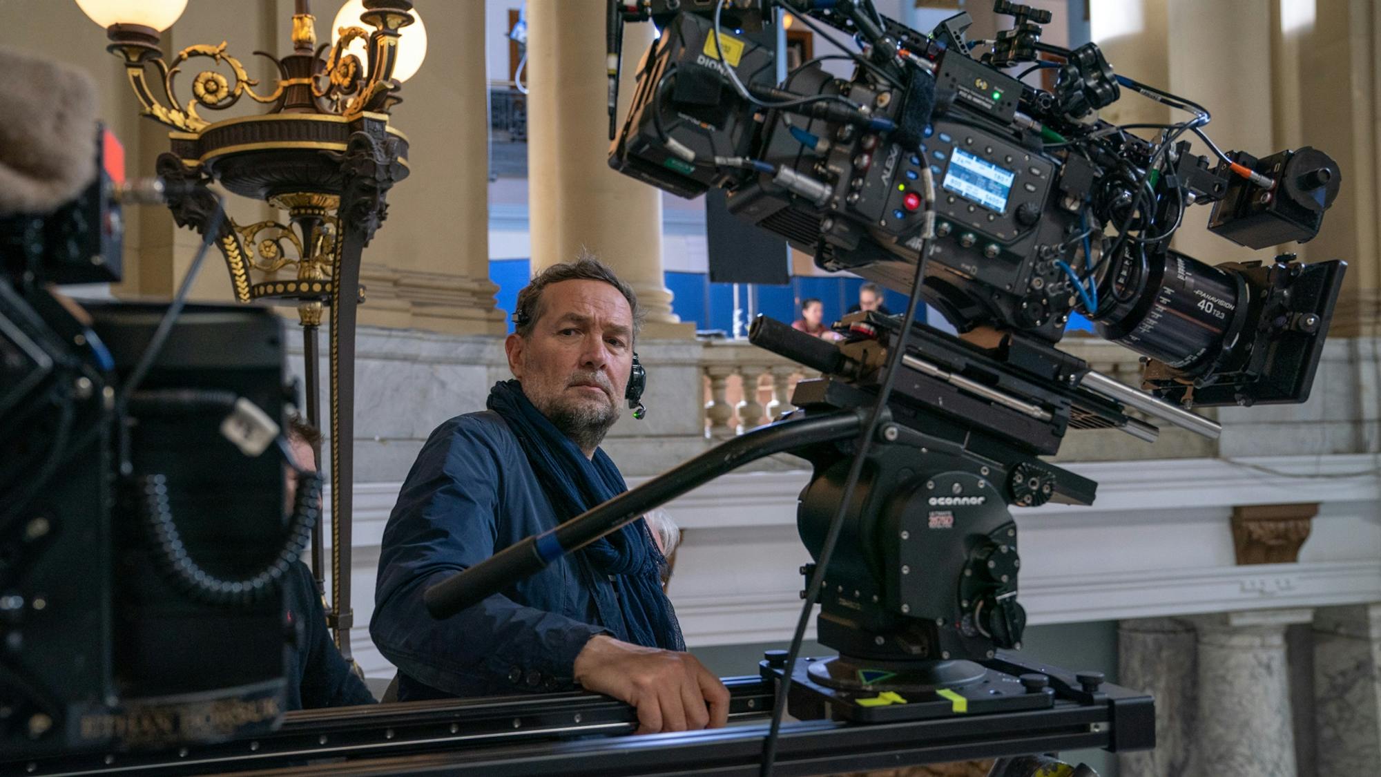 Papamichael is pictured in the courthouse set for The Trial of the Chicago 7. Behind him are marble pillars and an ornate, gilt lamp. He holds onto a camera rig with one hand, leaning back and looking toward us.