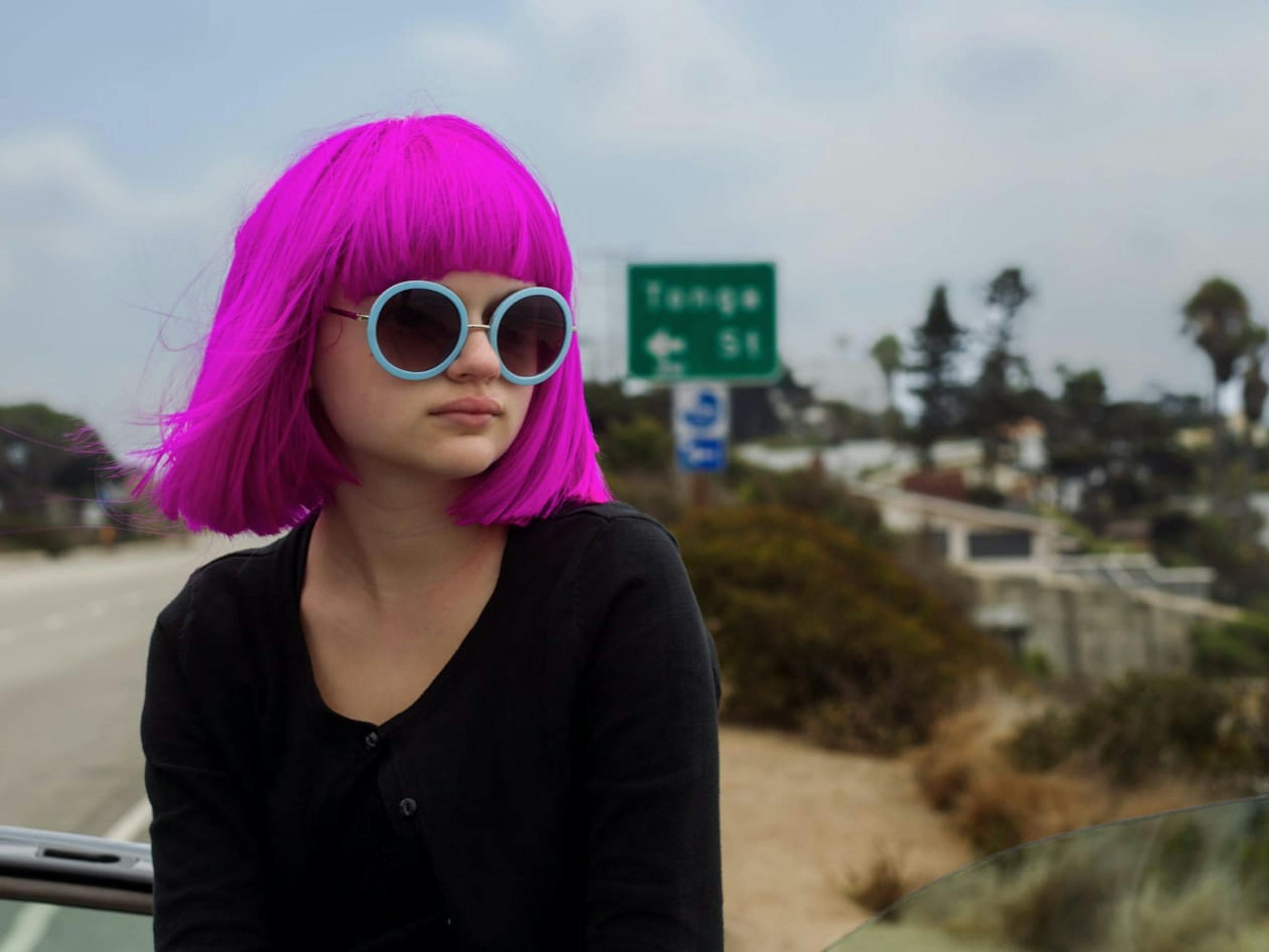 Grace Bloom (Joey King) in Wish I Was Here (2014) wears a pink bob, circular glasses, and a black top. In the background is a green road sign and trees.