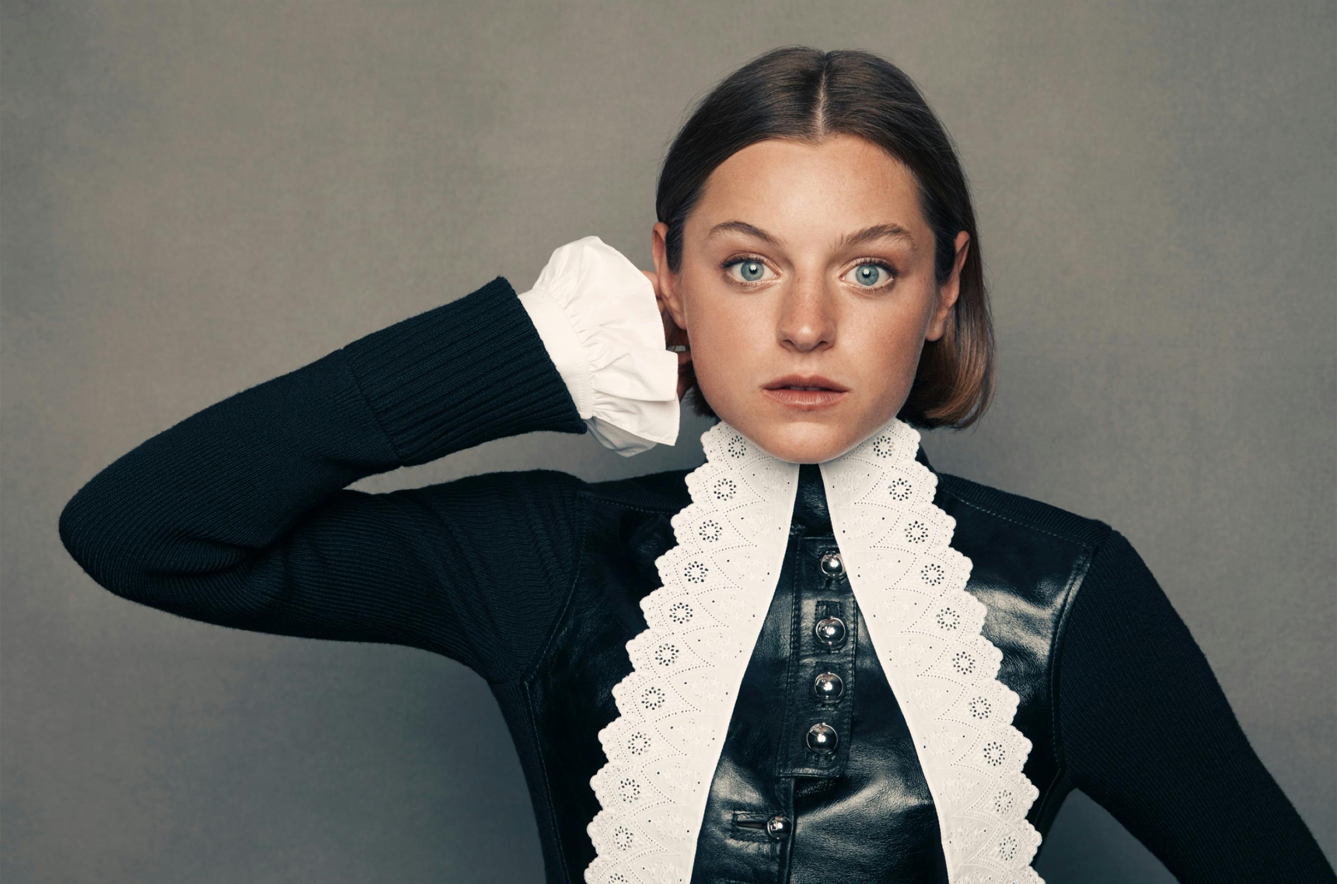 A portrait of Emma Corrin set against a slate-gray background. She wears a striking black leather and lace outfit with long sleeves and a high neckline. She has short cropped brown hair and her blue eyes pierce through the shot.