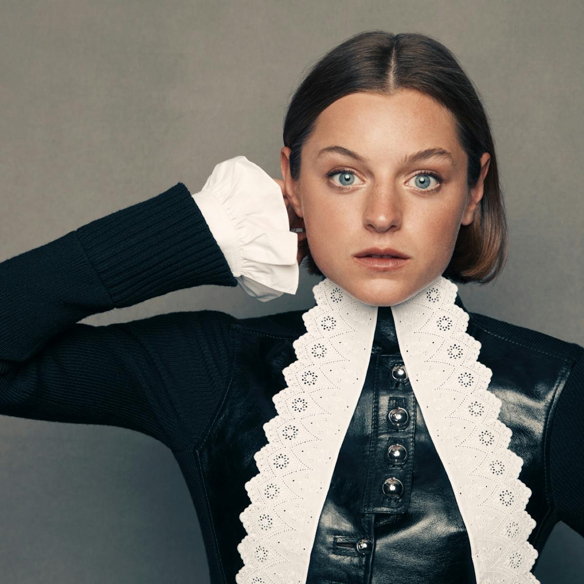 A portrait of Emma Corrin set against a slate-gray background. She wears a striking black leather and lace outfit with long sleeves and a high neckline. She has short cropped brown hair and her blue eyes pierce through the shot.