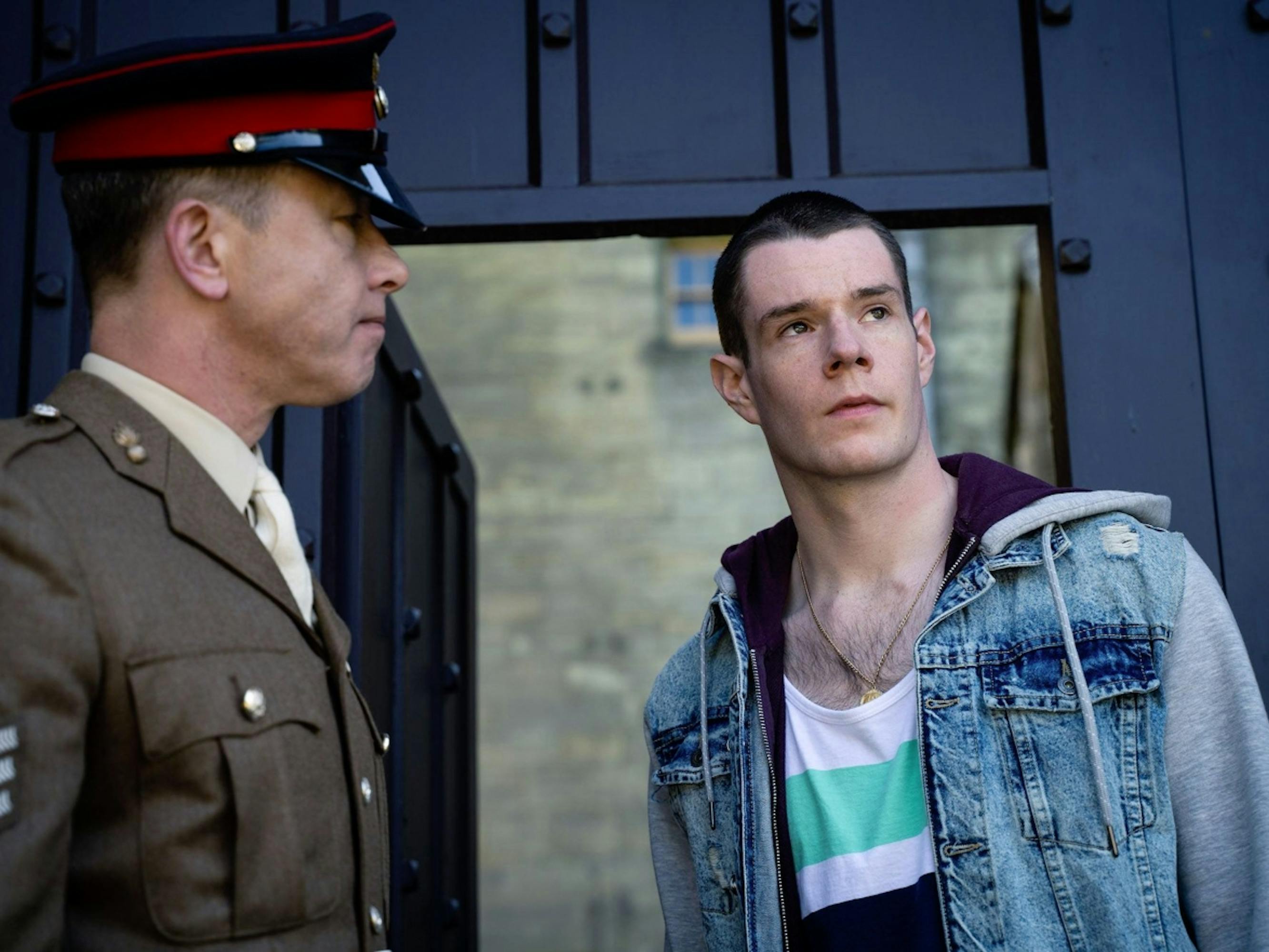 Adam Groff wears a tshirt and jean jacket and looks off into the distance. Beside him stands an army official in full dress.