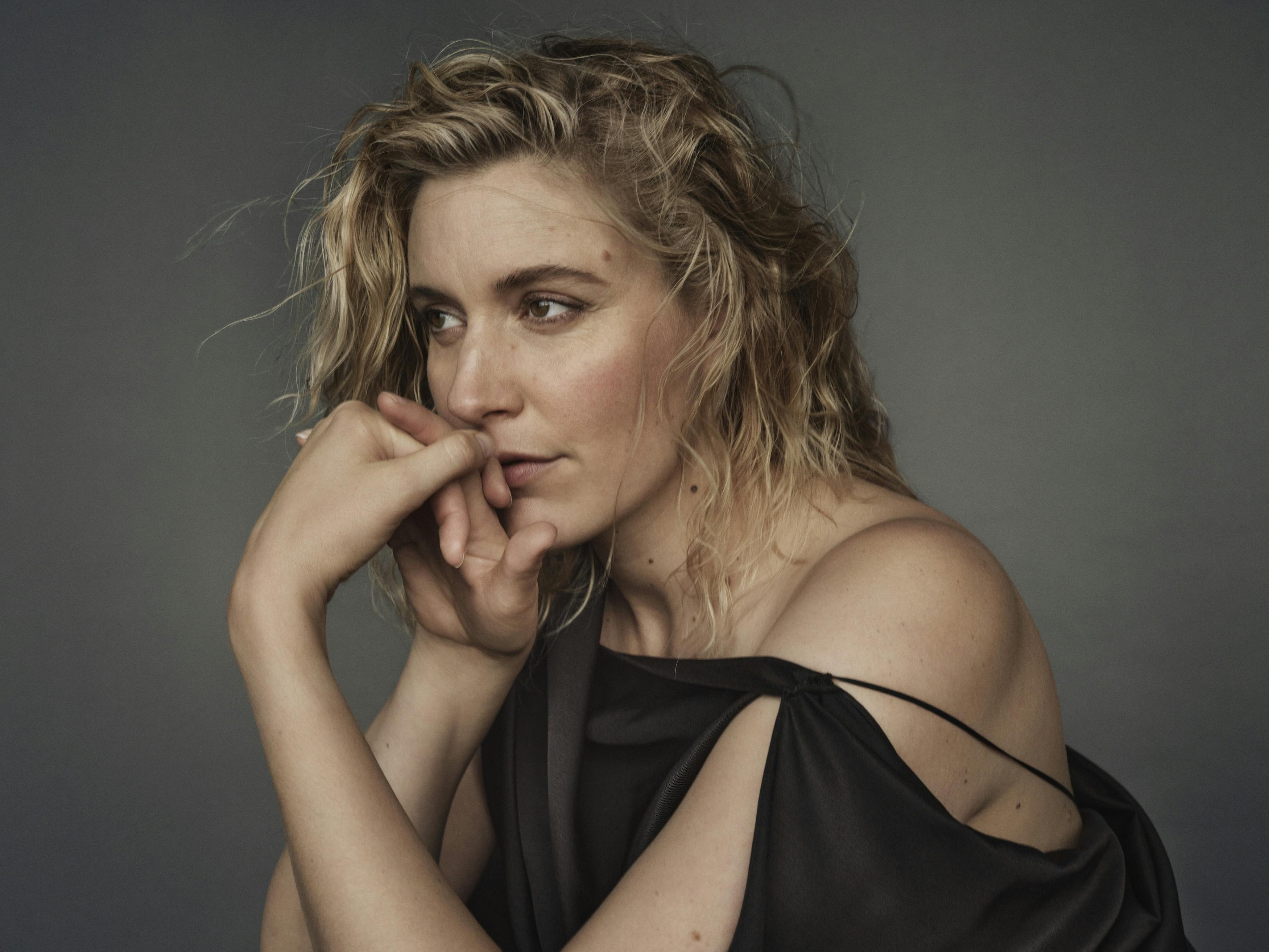 Greta Gerwig wears a black strapless top and rests her chin on her folded hands.