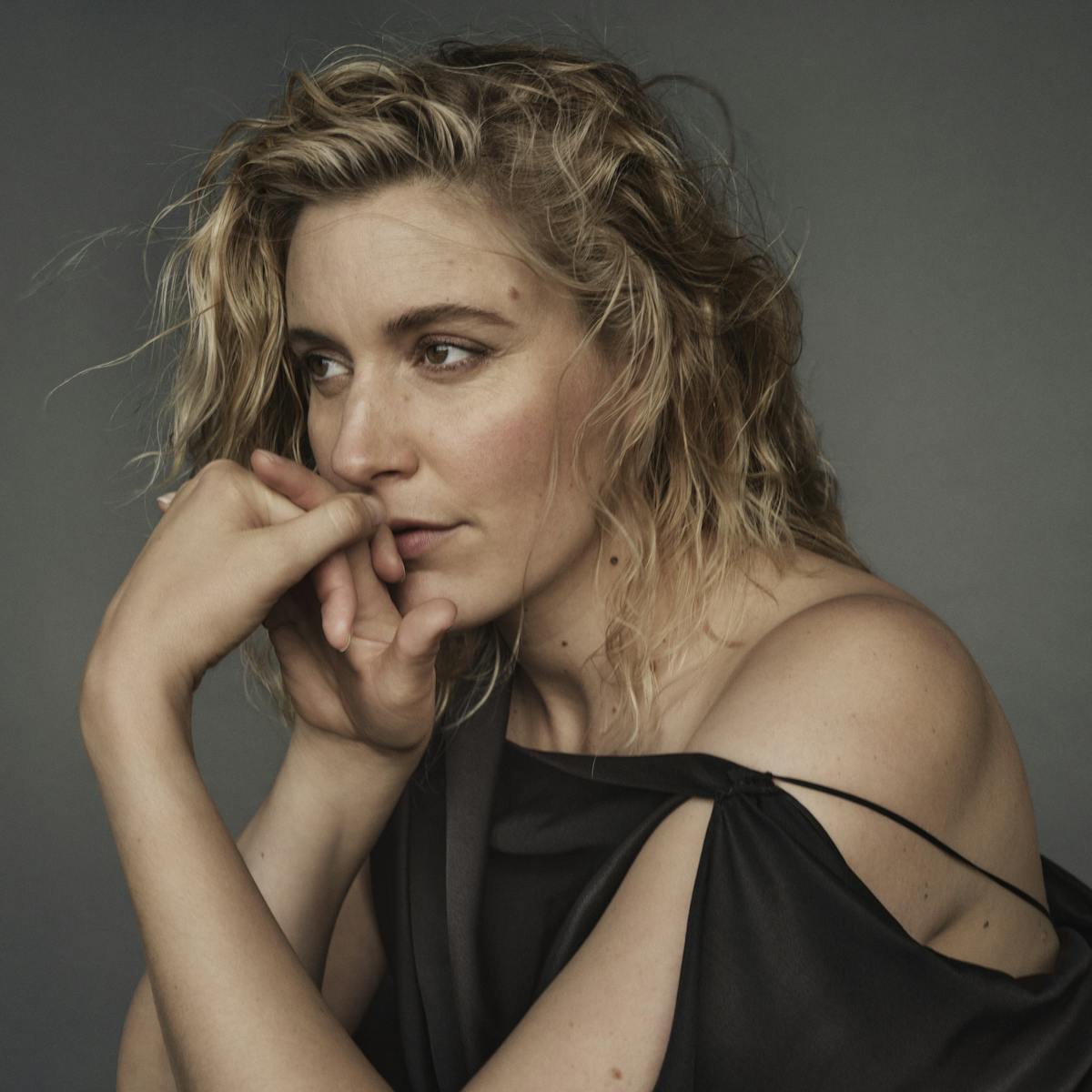 Greta Gerwig wears a black strapless top and rests her chin on her folded hands.