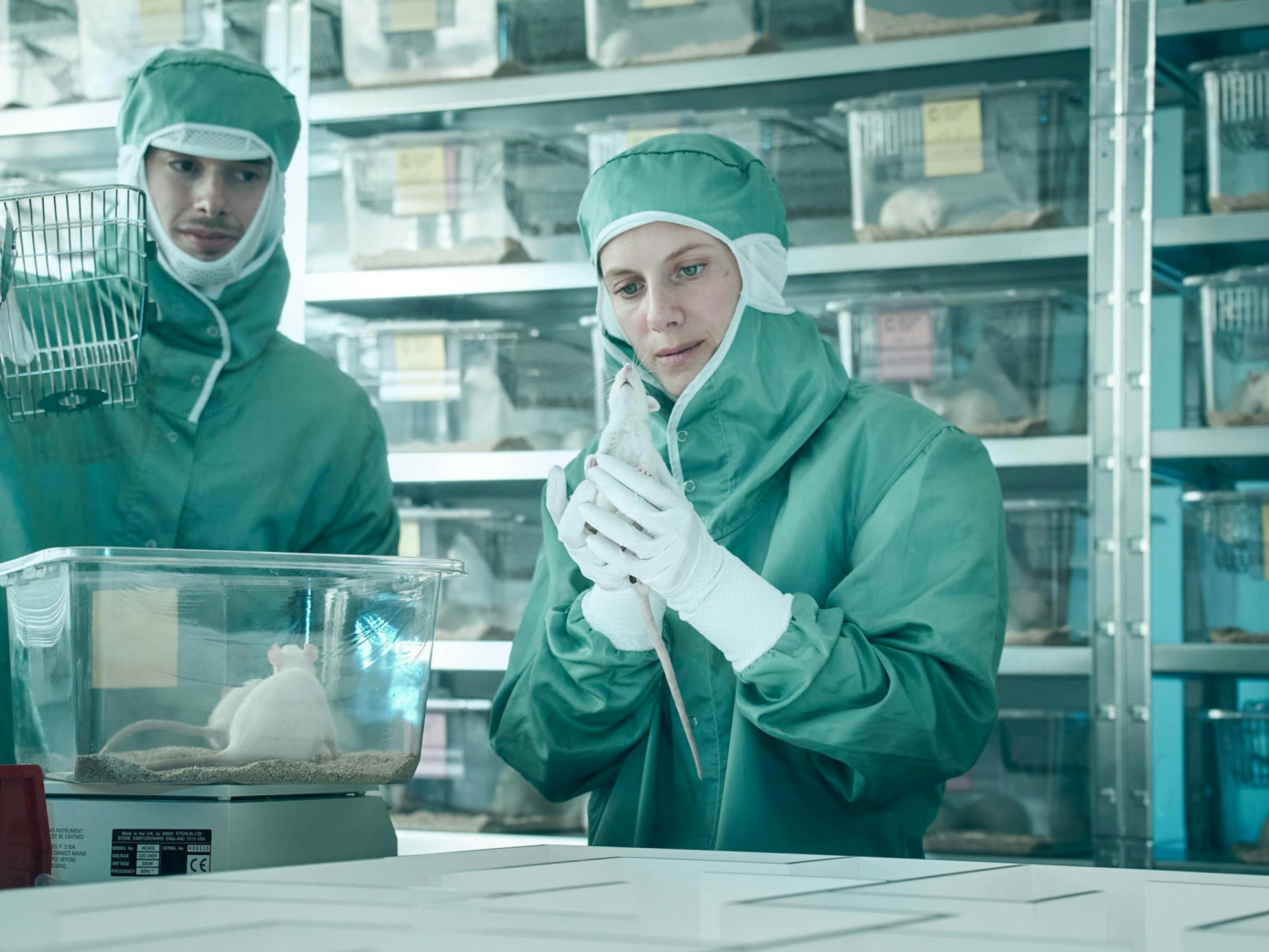 In what appears to be a flashback based on the lighting, Laurent and a man stand in a lab, dressed head to toe in green medical garb. She holds a white mouse in her hand, and to her right and behind her are many other white mice in clear boxes.