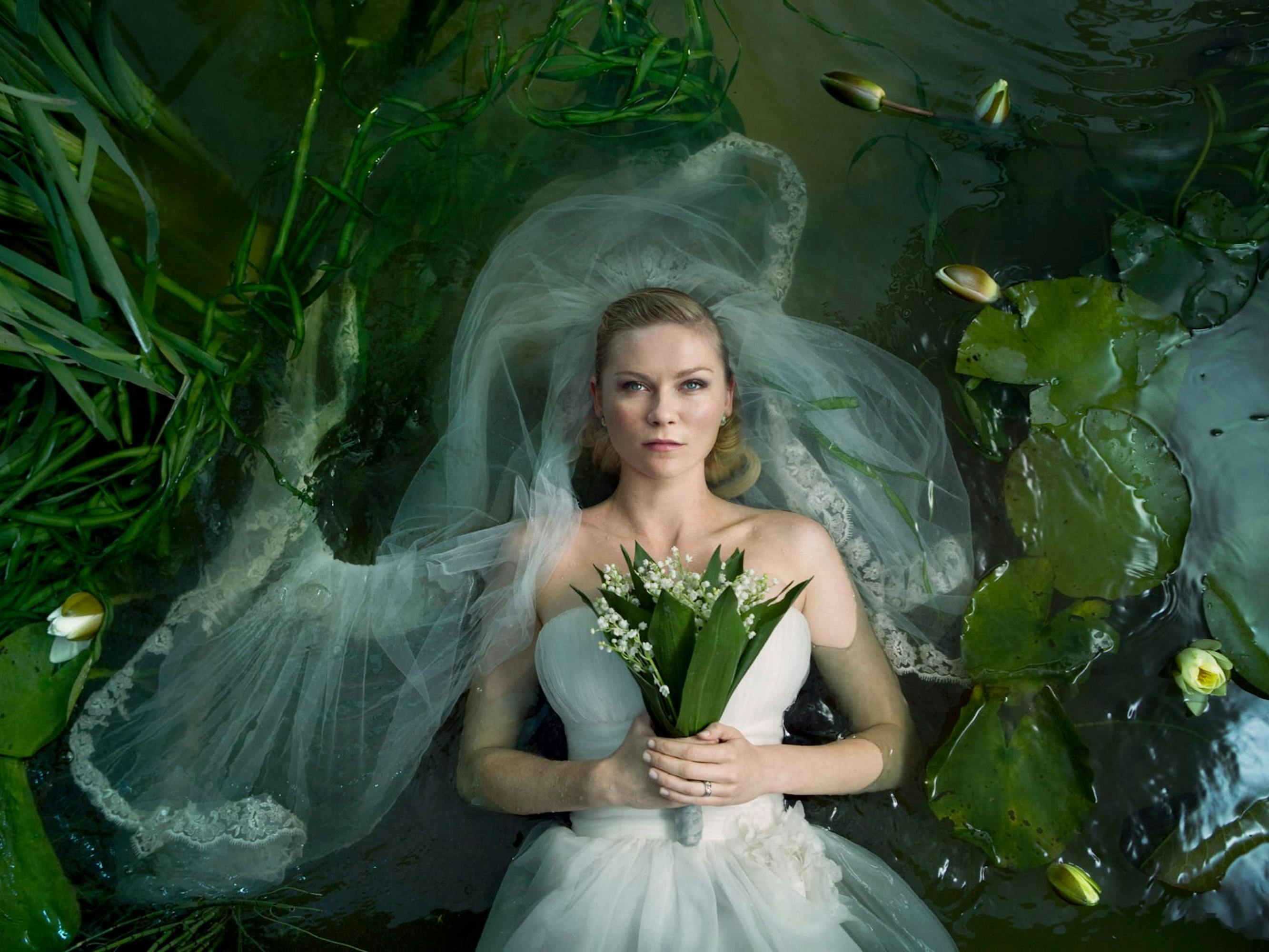 Justine (Kirsten Dunst) wears a wedding dress and holds a bouquet to her chest. She’s surrounded by lilypads and other greenery in a murky pond.