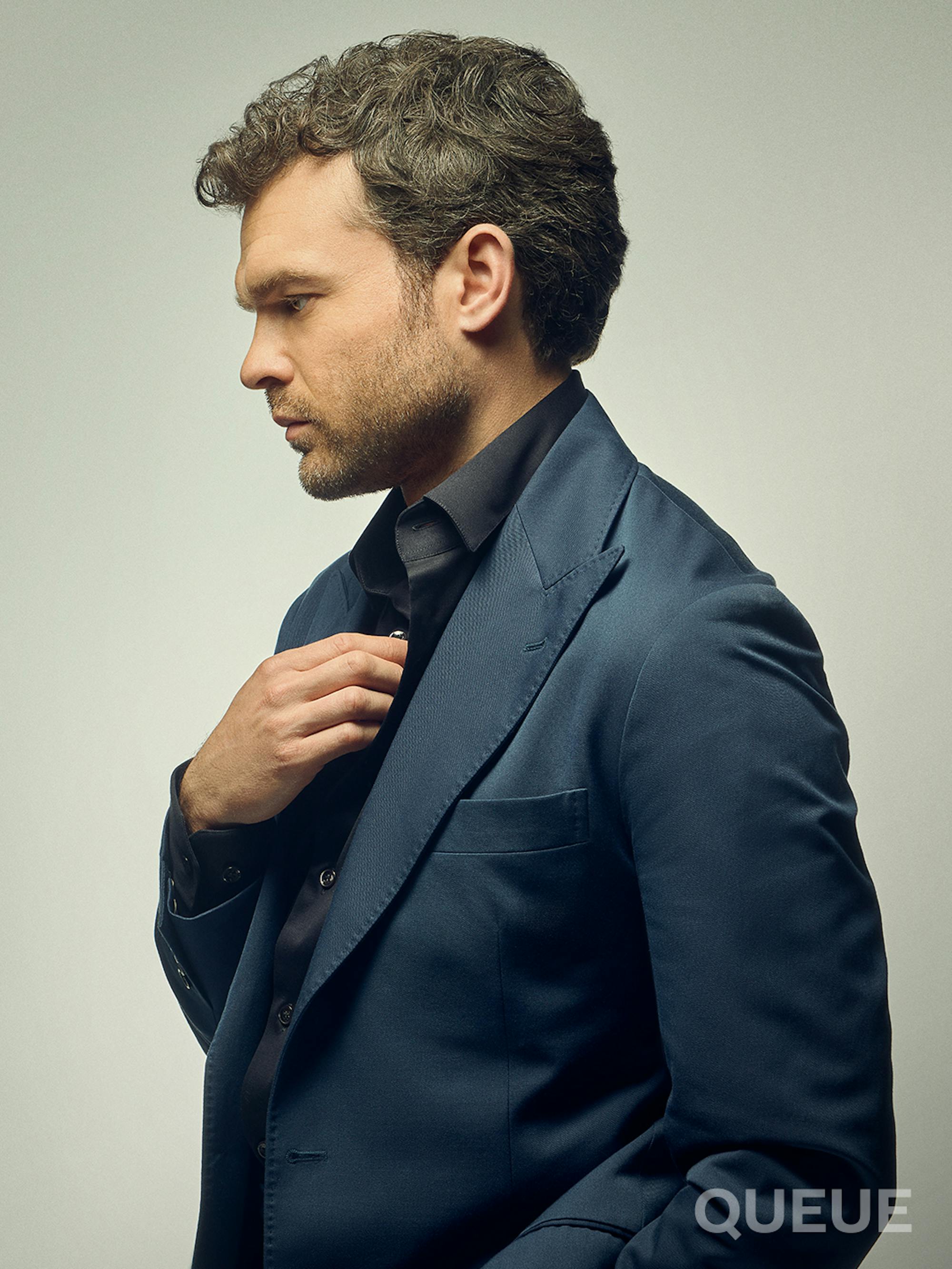 Alden Ehrenreich stands in profile and wears a navy suit.