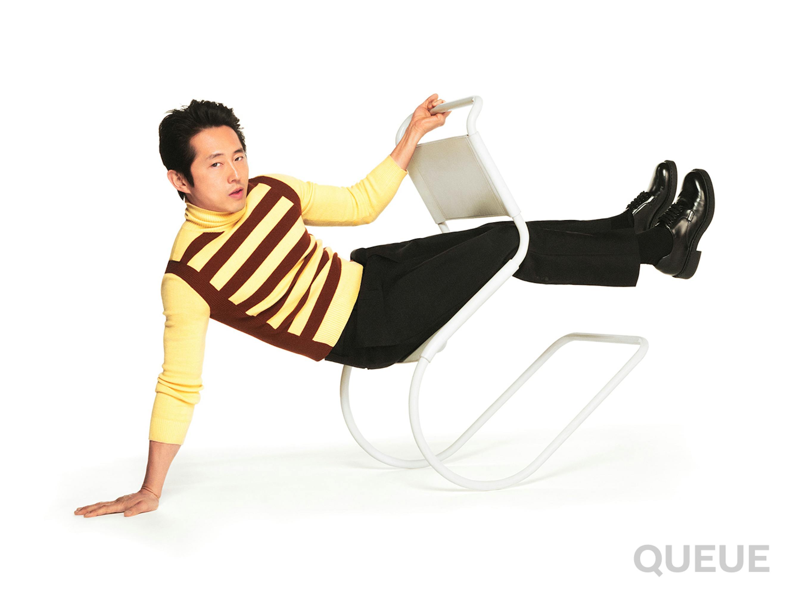 Steven Yeun leans back in a white chair. He wears a yellow and maroon striped shirt and black pants.