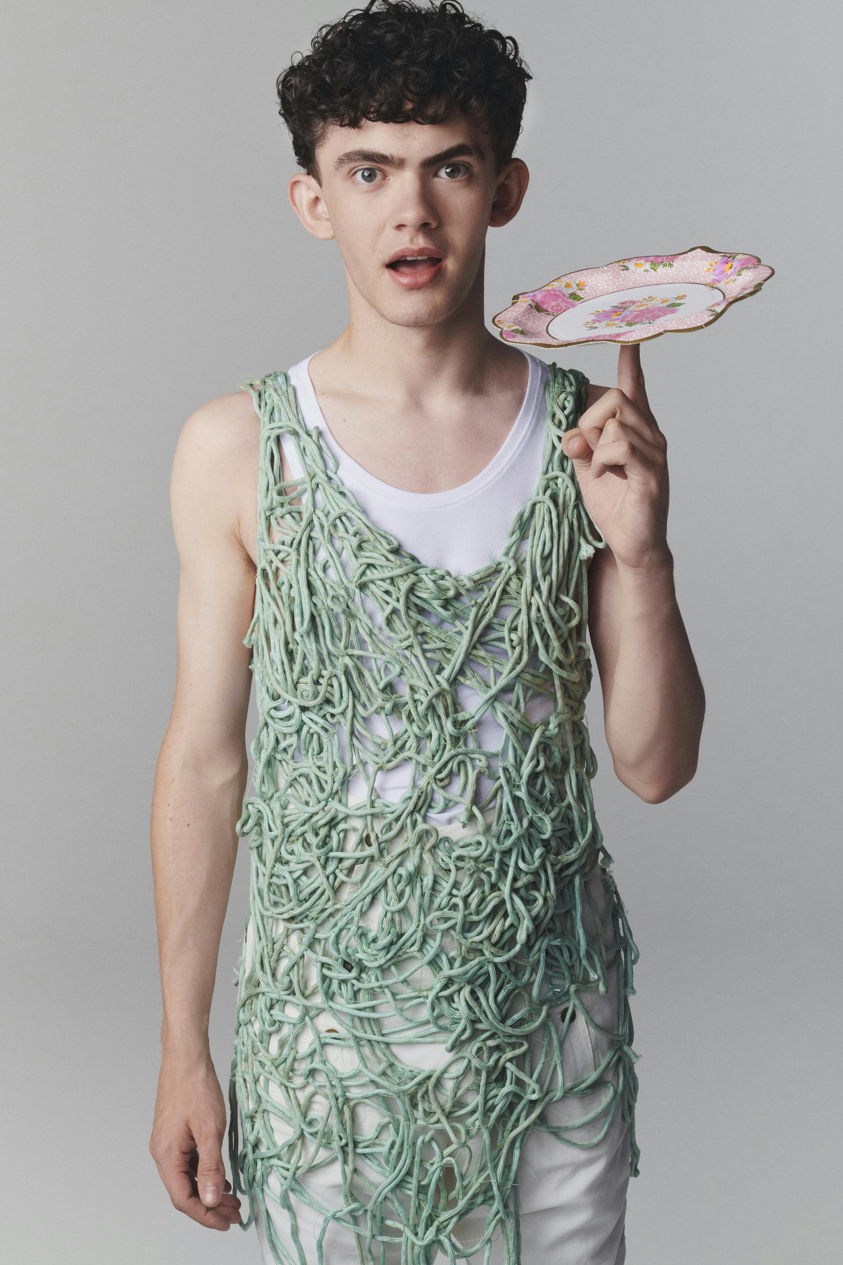 Joe Locke wears a green rope vest and white tank top, and spins a pink floral plate on his finger. 