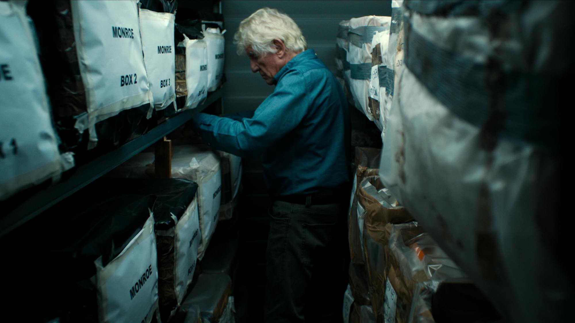 Anthony Summers wears a jean top and looks through boxes labeled 'Monroe Boxs' in what seems like a storage unit.
