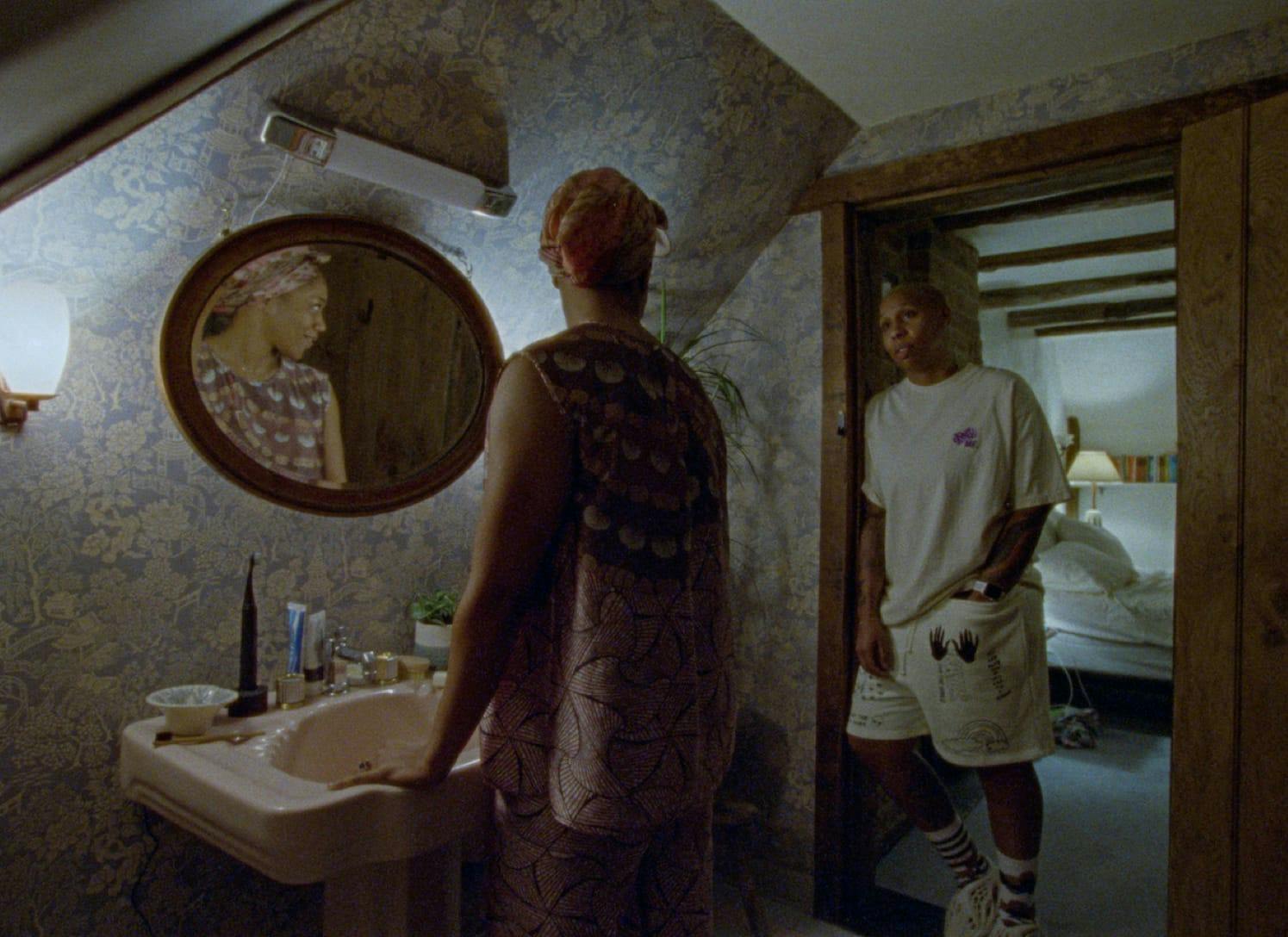 Denise (Lena Waithe) and Alicia (Naomi Ackie) stand in their bathroom. The light makes it look like nighttime. Alicia stands at the sink and wears patterned pajamas, and Denise wears all white.