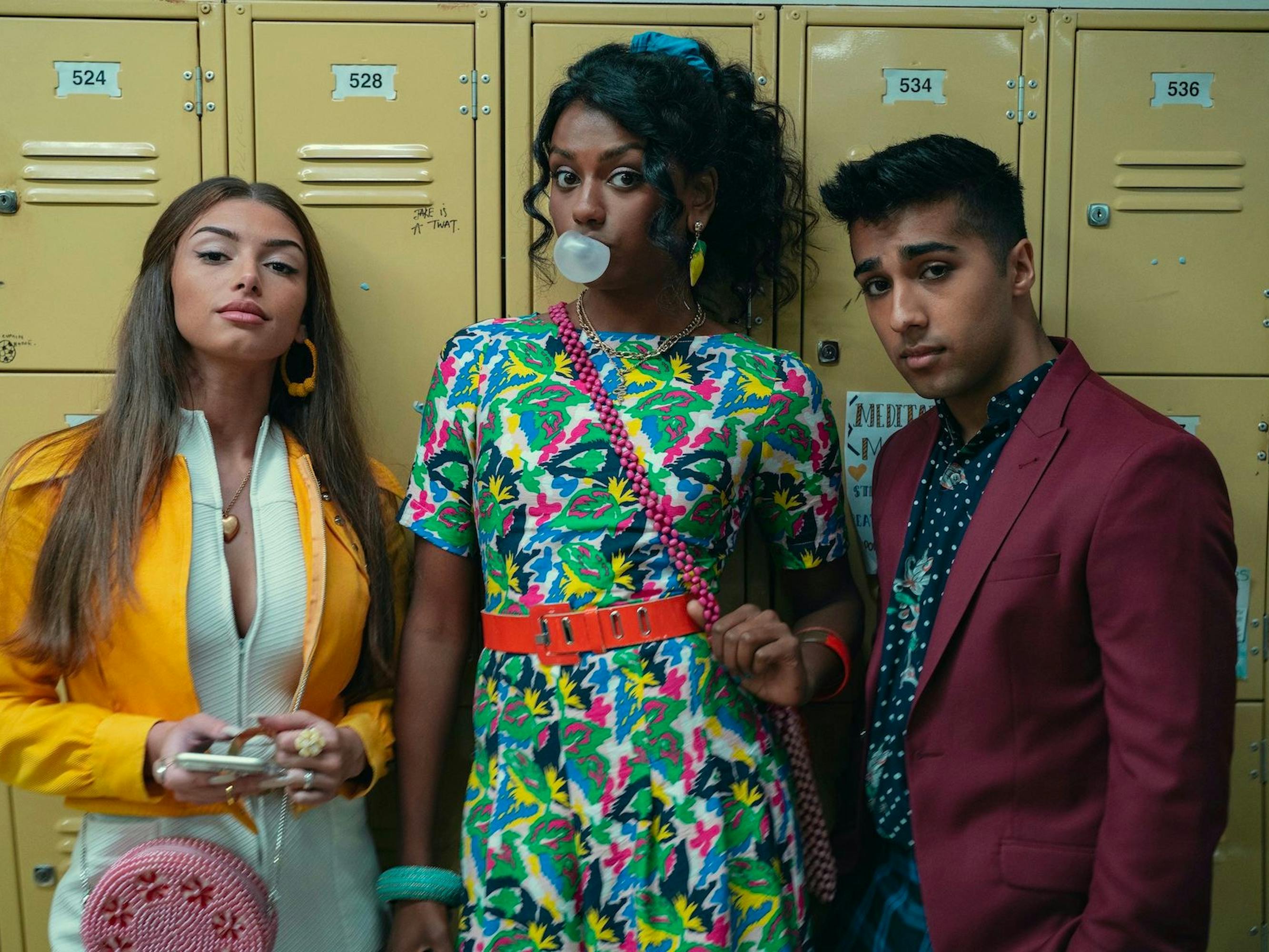 Ruby Matthews (Mimi Keene), Olivia Hanan (Simone Ashley), and Anwar (Chaneil Kular) stand against the tan lockers look fierce and scary. Ashley blows a customary bubble.