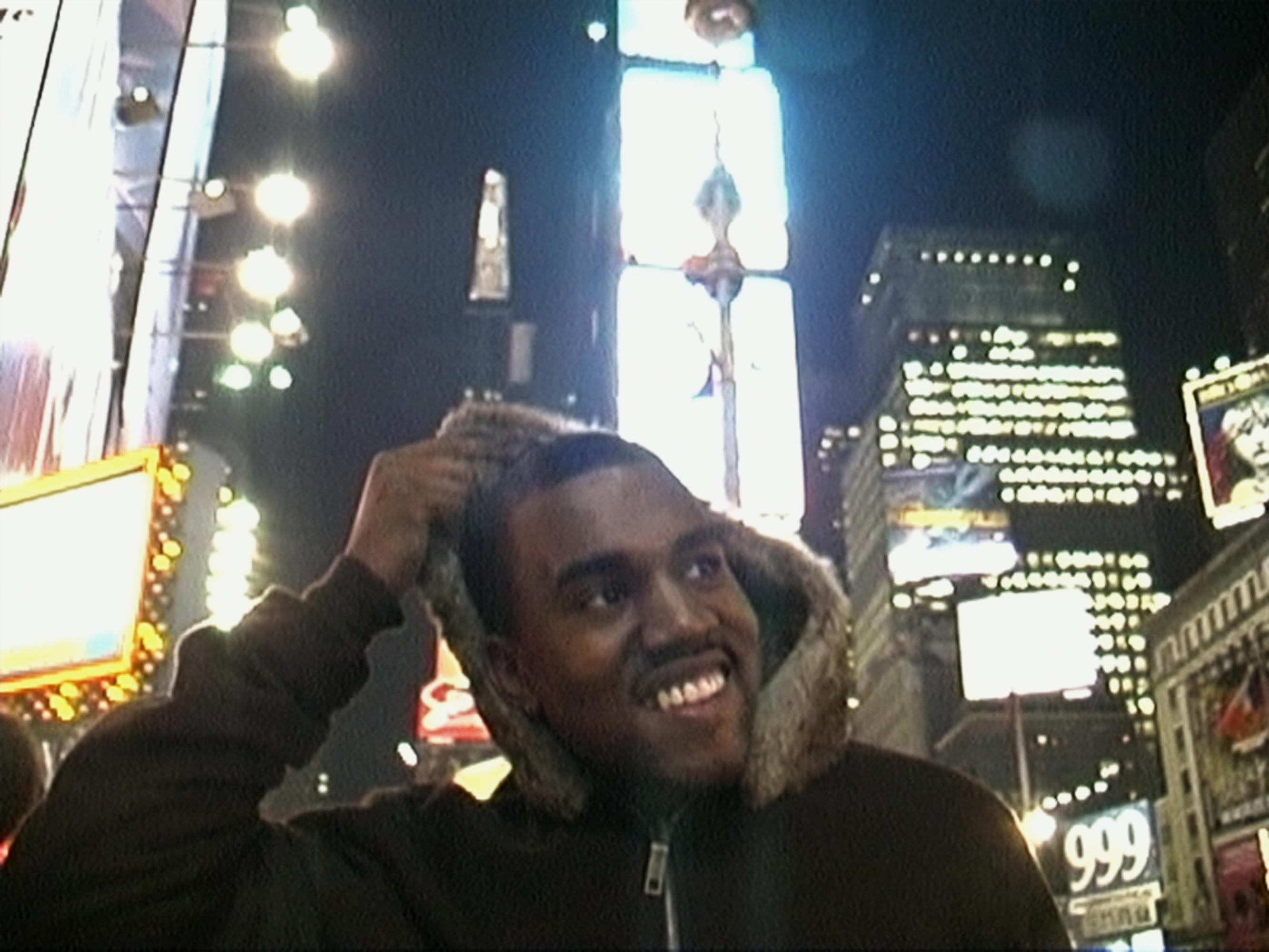 Kanye West wears a brown jacket with a furry hood. Behind him are illuminated buildings.