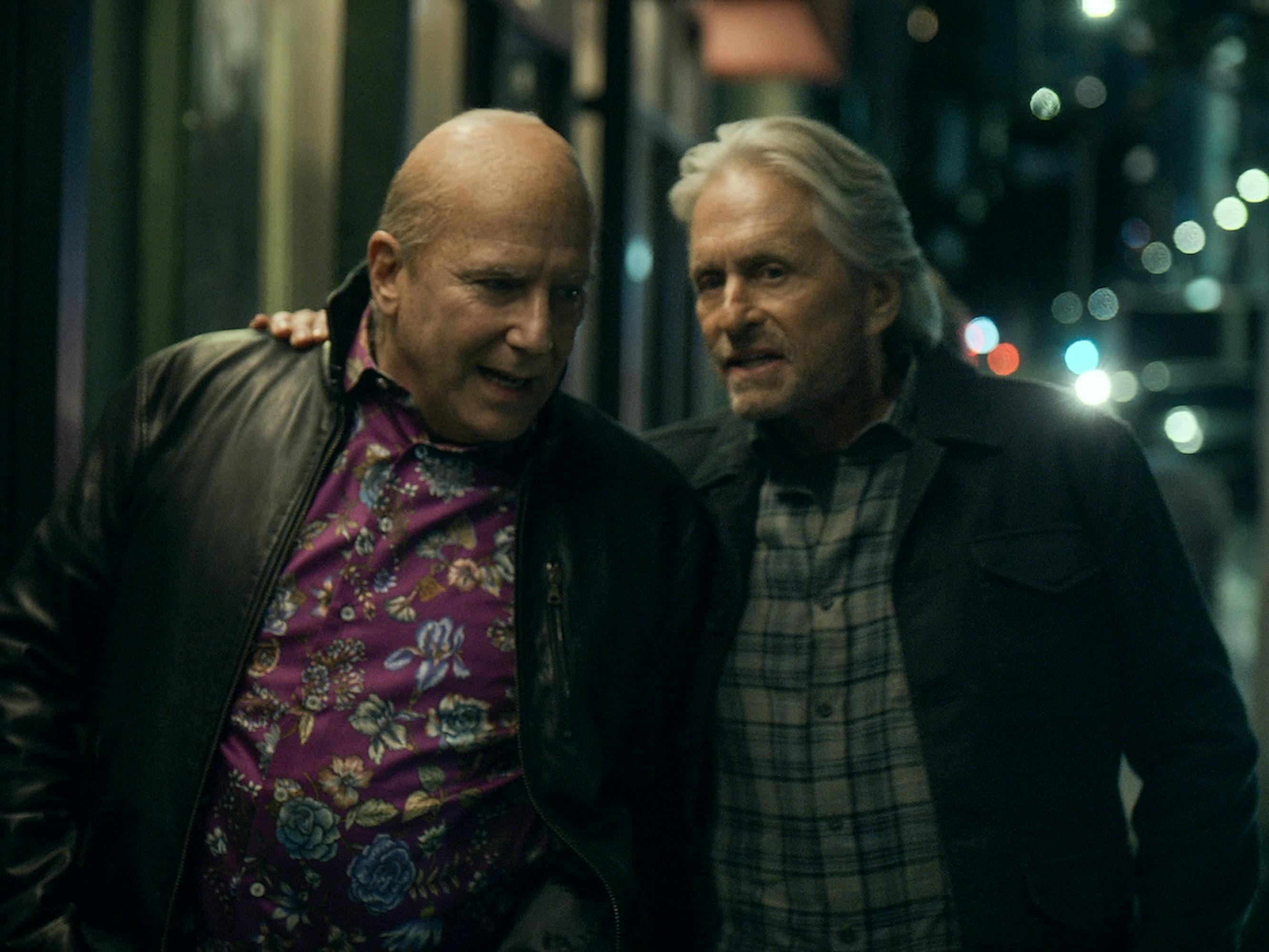Martin (Paul Reiser) and Sandy Kominsky (Michael Douglas) walk together on a sidewalk. Reiser wears a purple floral shirt and Douglas wears a plaid one.