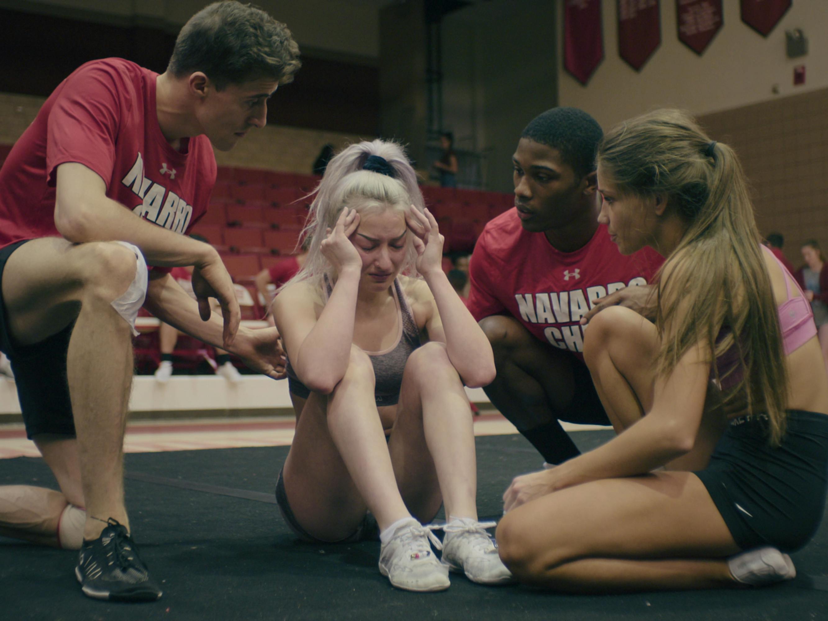 Dillon Brandt, Lexi Brumback, La'Darius Marshall and Morgan Simianer huddle together on a black mat. Lexi looks upset and the other three comfort her. The two men wear red shirts and black shorts. Lexi wears white sneakers and a grey sports bra, and Morgan wears black shorts and a pink sports bra.