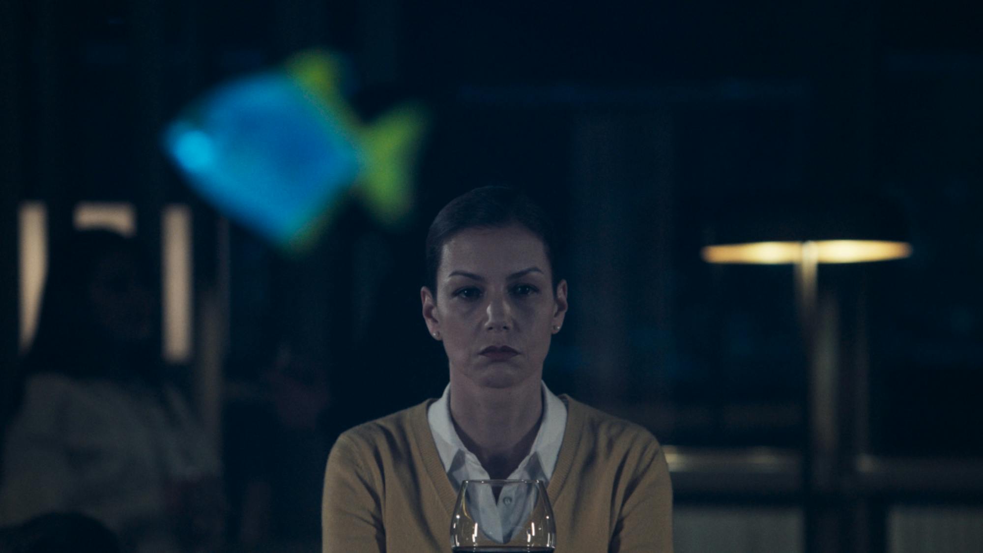 Peri looks stoic sitting behind a fish tank with a glass of wine.