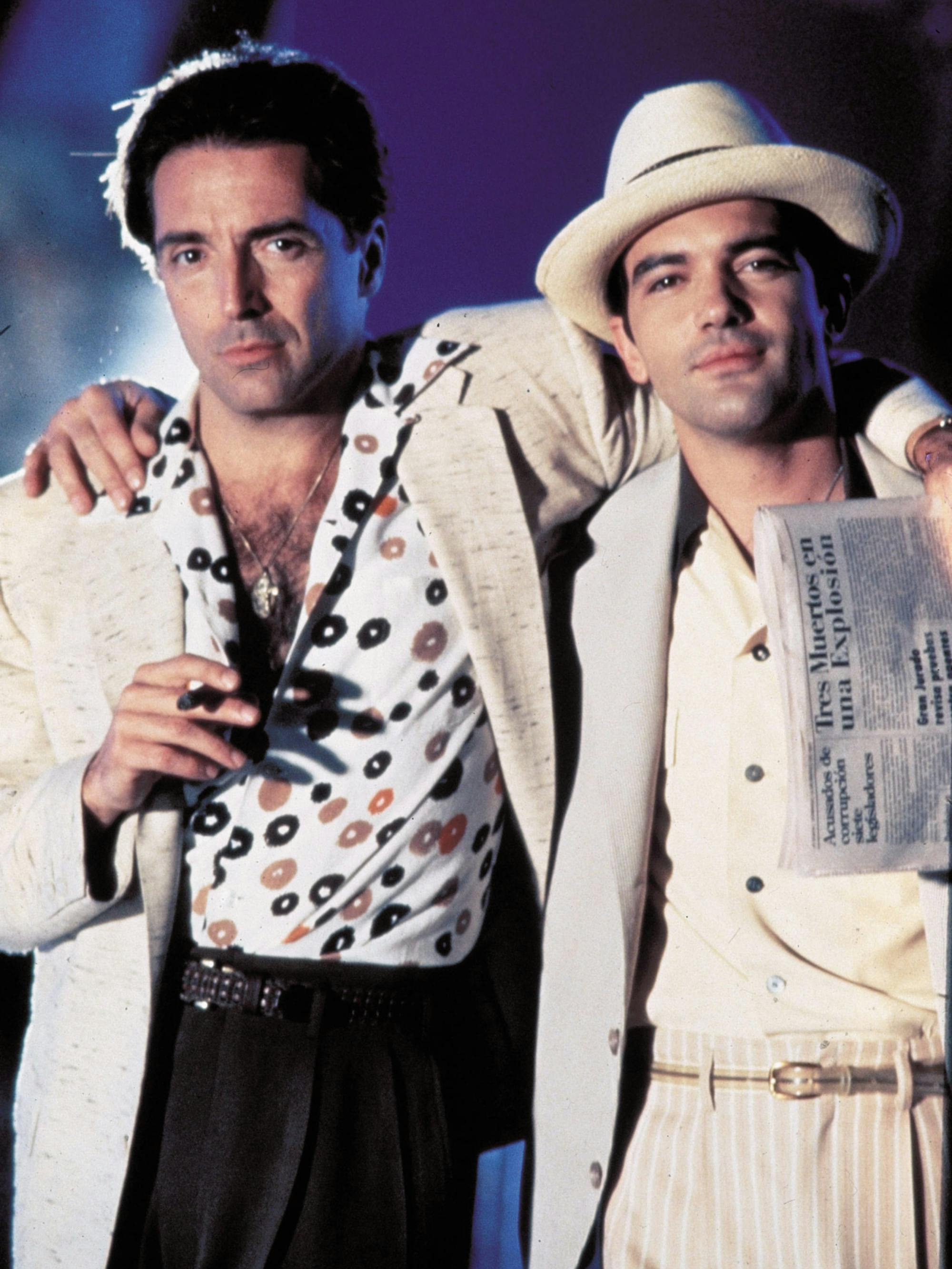 Armand Assante and Antonio Banderas stand with their arms around each others shoulders in a still from The Mambo Kings. Assante’s white button-down shirt with polka dots in black, orange and tan is unbuttoned far down his chest. Banderas’ cream-colored hat tops off his all white suit. 