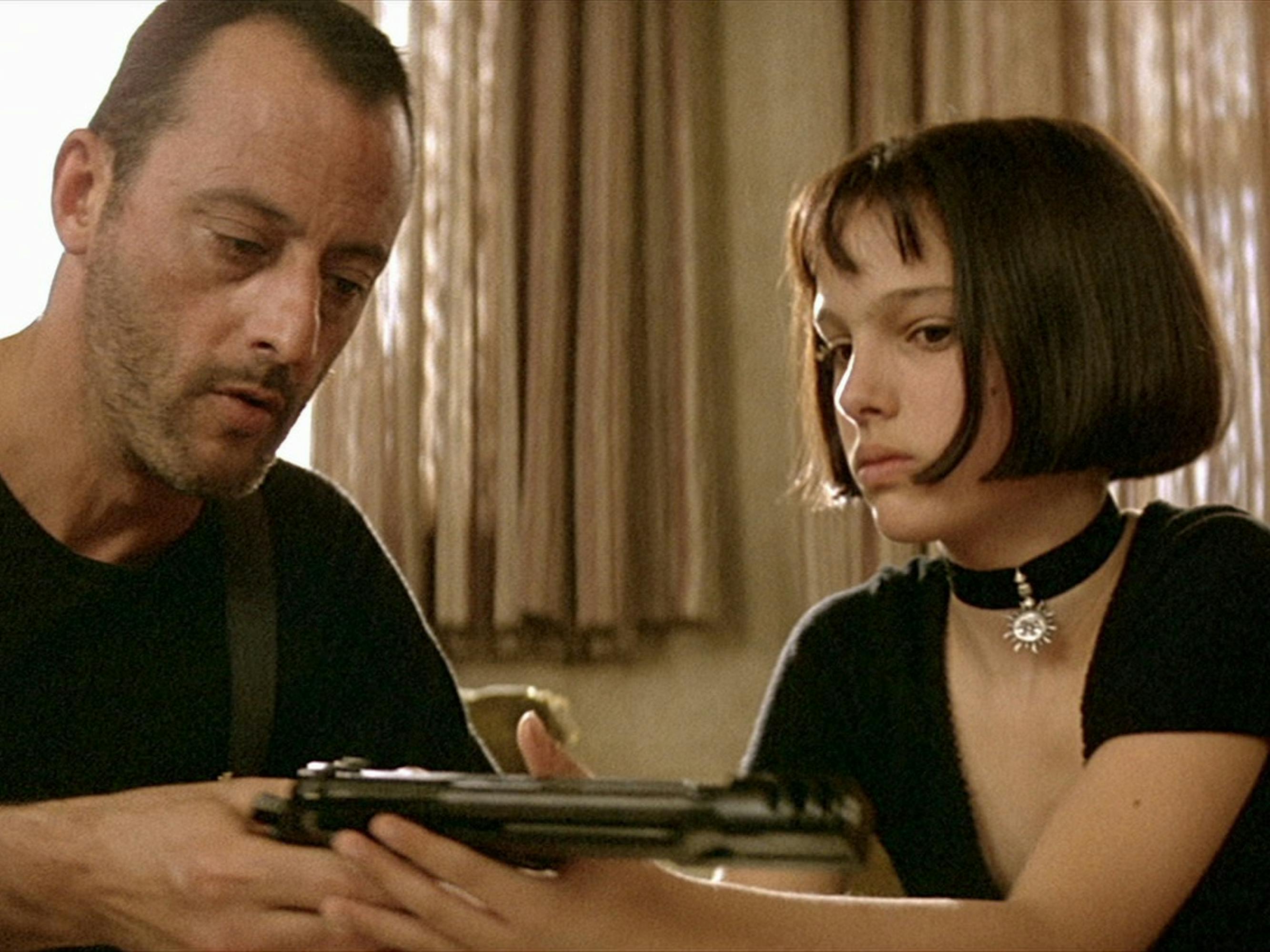 Léon (Jean Reno) and Mathilda (Natalie Portman) sit in a beige room looking at a gun.