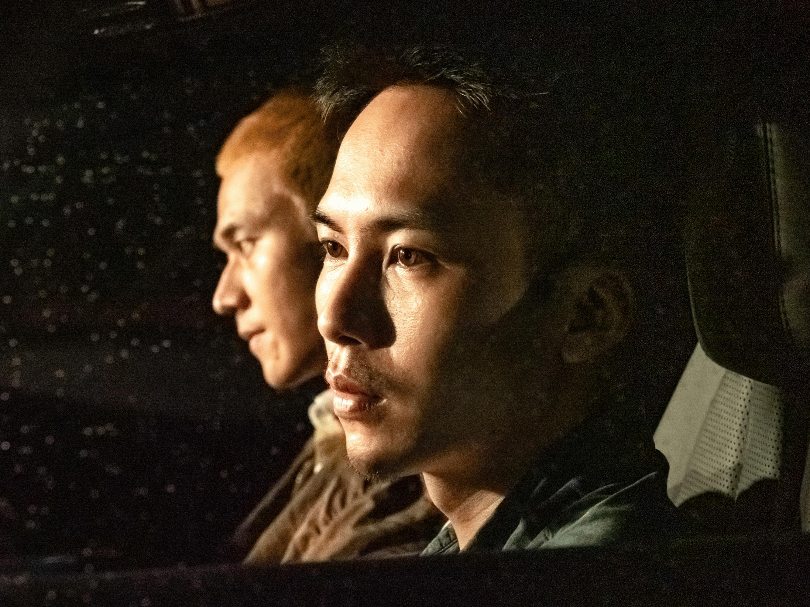 Radish and A-ho sit in a dark car in this still from the film. Their faces are illuminated, and the two young men look in different directions. Radish’s bleached hair stands out in the darkness around him.