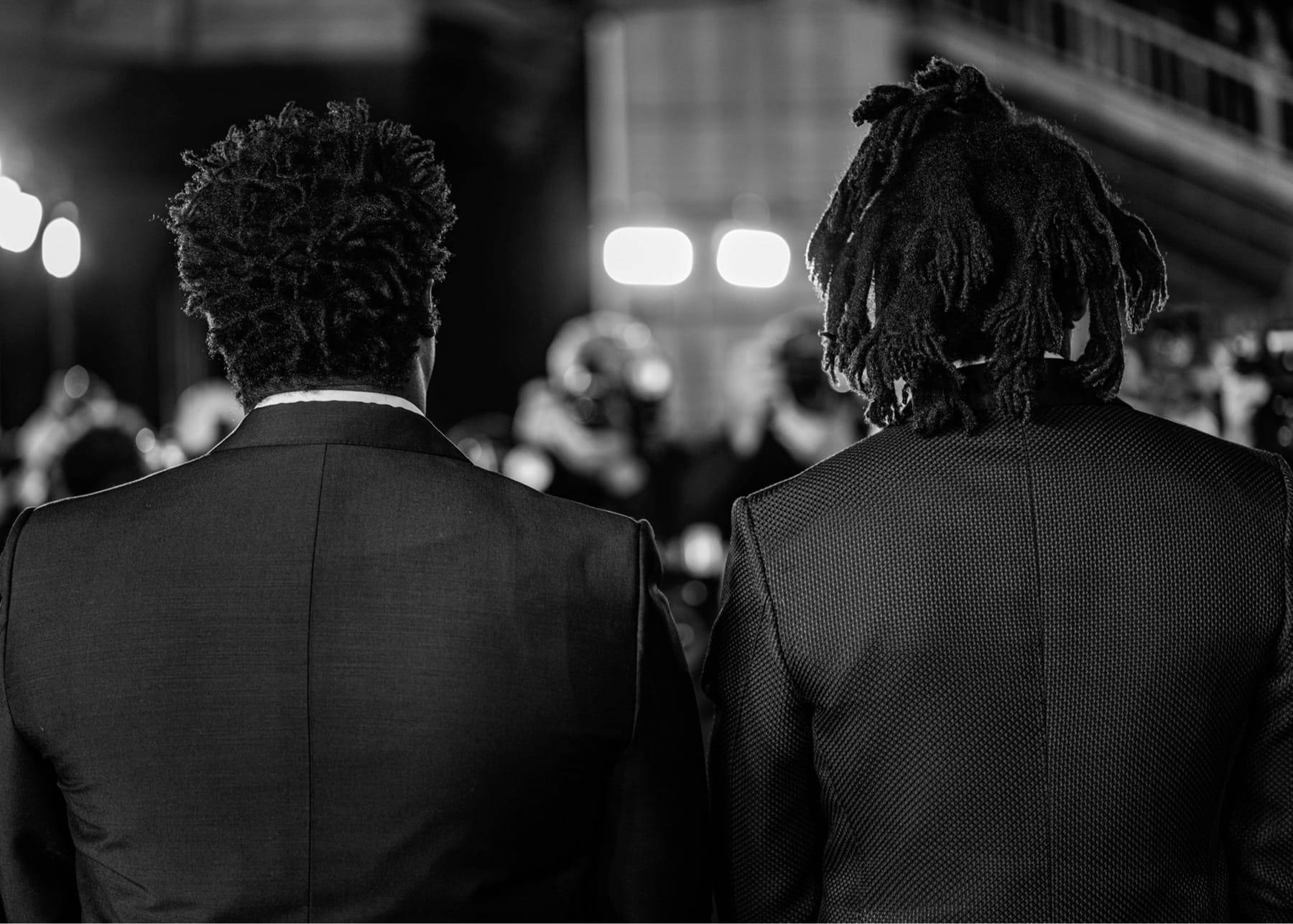 Jeymes Samuel and Jay-Z stand shoulder to shoulder in dark blazers in this close-up, from-behind, black-and-white shot.