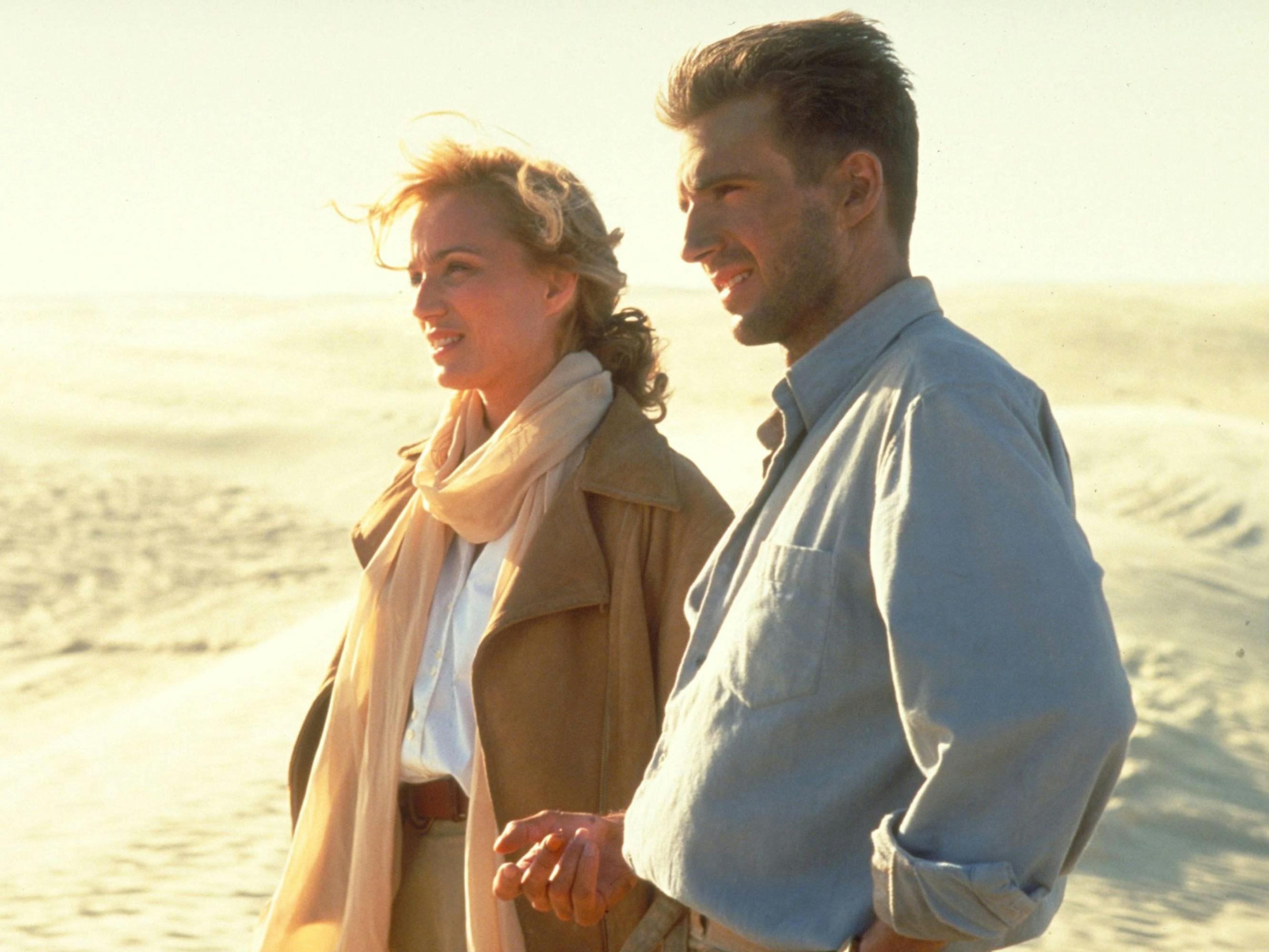 Kristin Scott Thomas and Ralph Fiennes stand together in a scene from The English Patient. Scott Thomas has a billowy tan scarf hung loosely around her neck. They smile, lit by the sun.