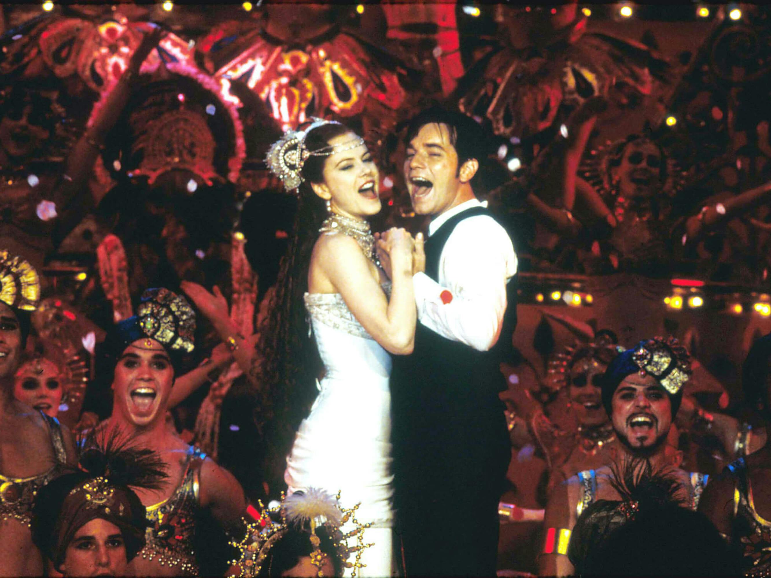 Satine (Nicole Kidman) and Christian (Ewan McGregor) in Moulin Rouge! He wears a suit, and she wears a white dress. People’s faces dot the background in this energetic shot.