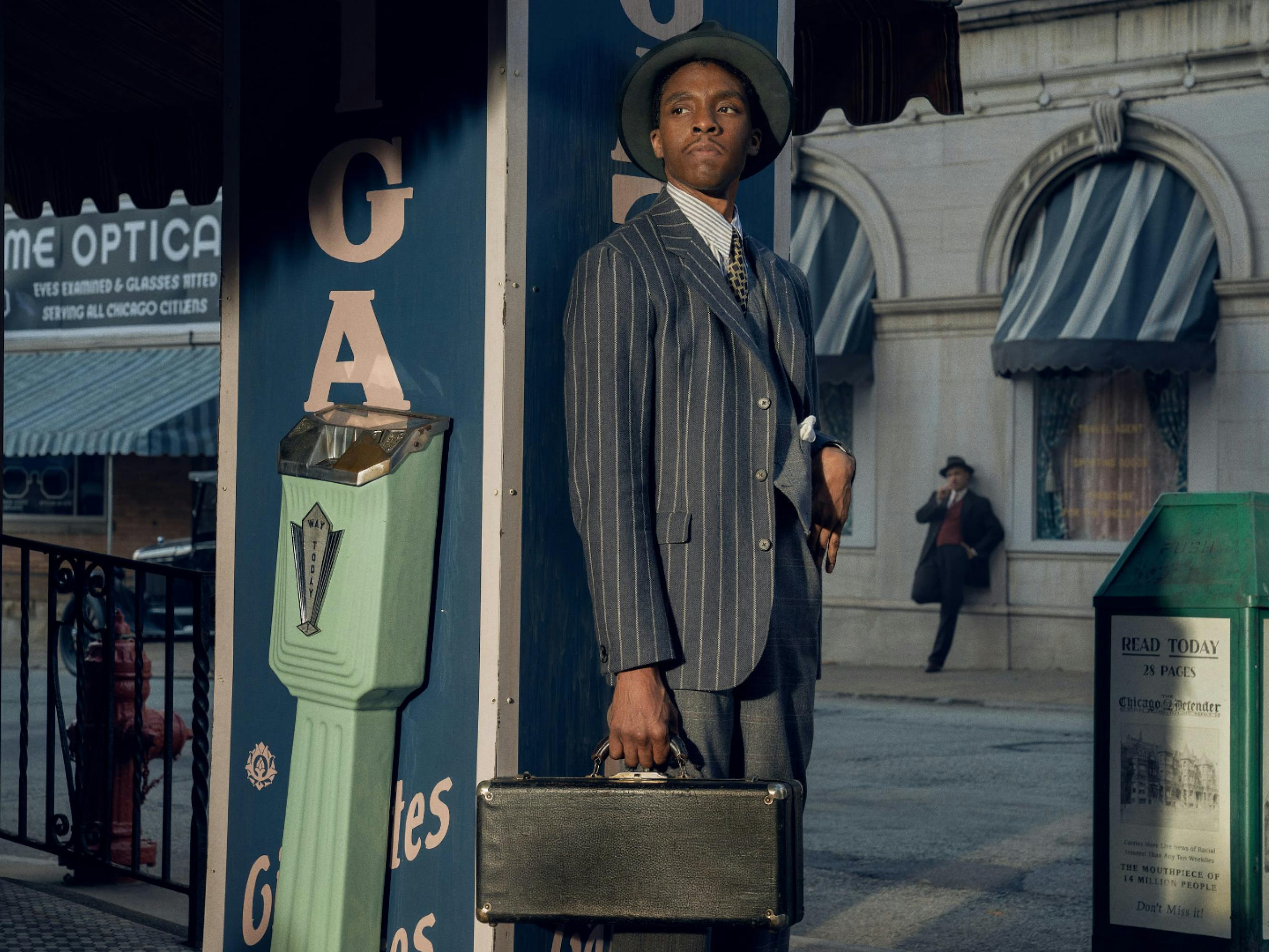 Boseman as Levee. He wears a striped blazer, grey hat, and carries a briefcase. In the background is another man leaning against a stone wall. There is an old green newspaper dispenser, and some striped awnings in the background. 