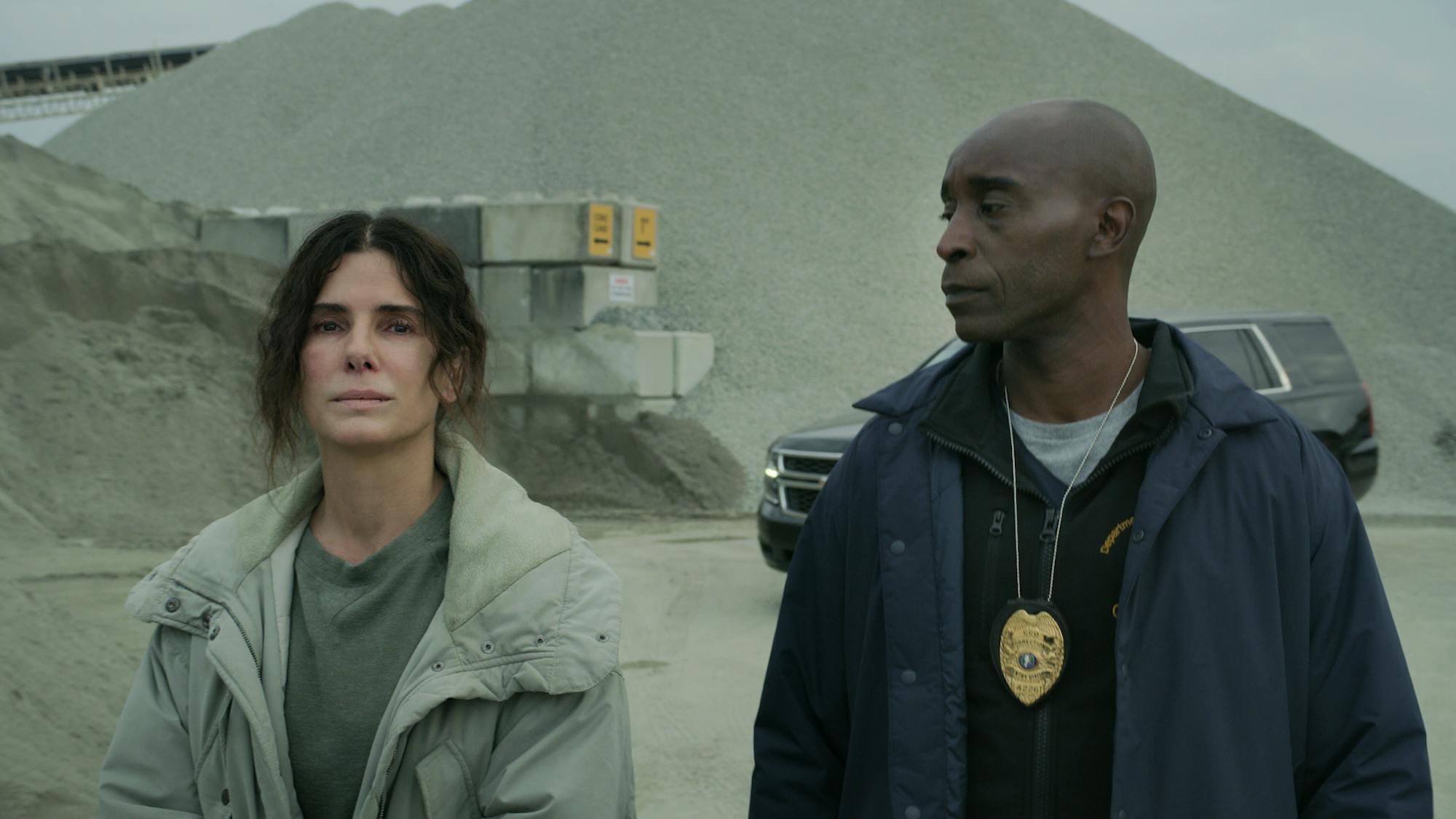 Ruth Slater (Sandra Bullock) and Vincent Cross (Rob Morgan) stand in a dusty lot. Vincent wears a navy jacket with a police badge, and Ruth wears an olive green shirt and light green jacket. 