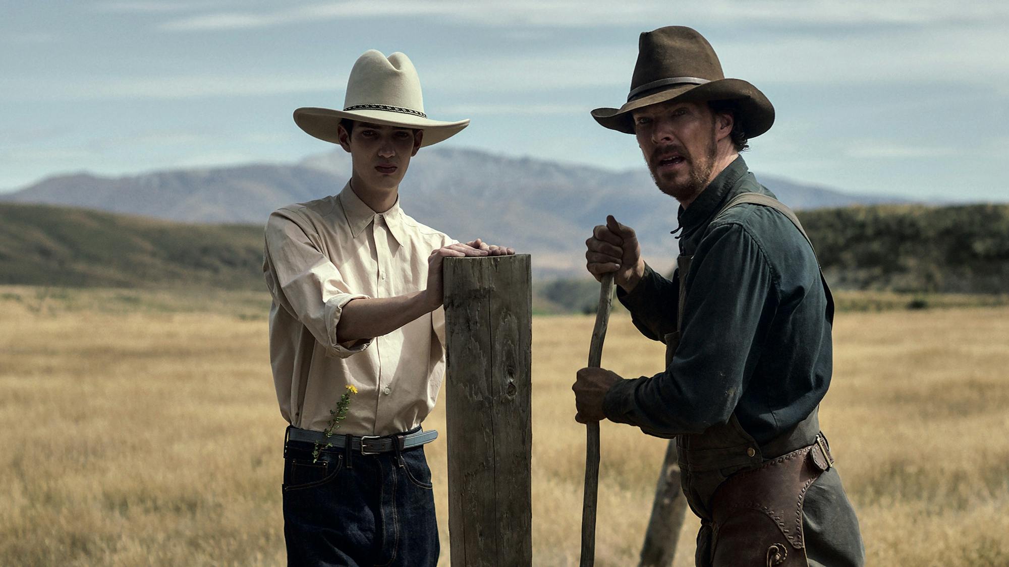 Peter Gordon (Kodi Smit-McPhee) and Phil Burbank (Benedict Cumberbatch) stand in a field. Behind them are green-blue mountains. They both wear wide brimmed hats and workshirts.