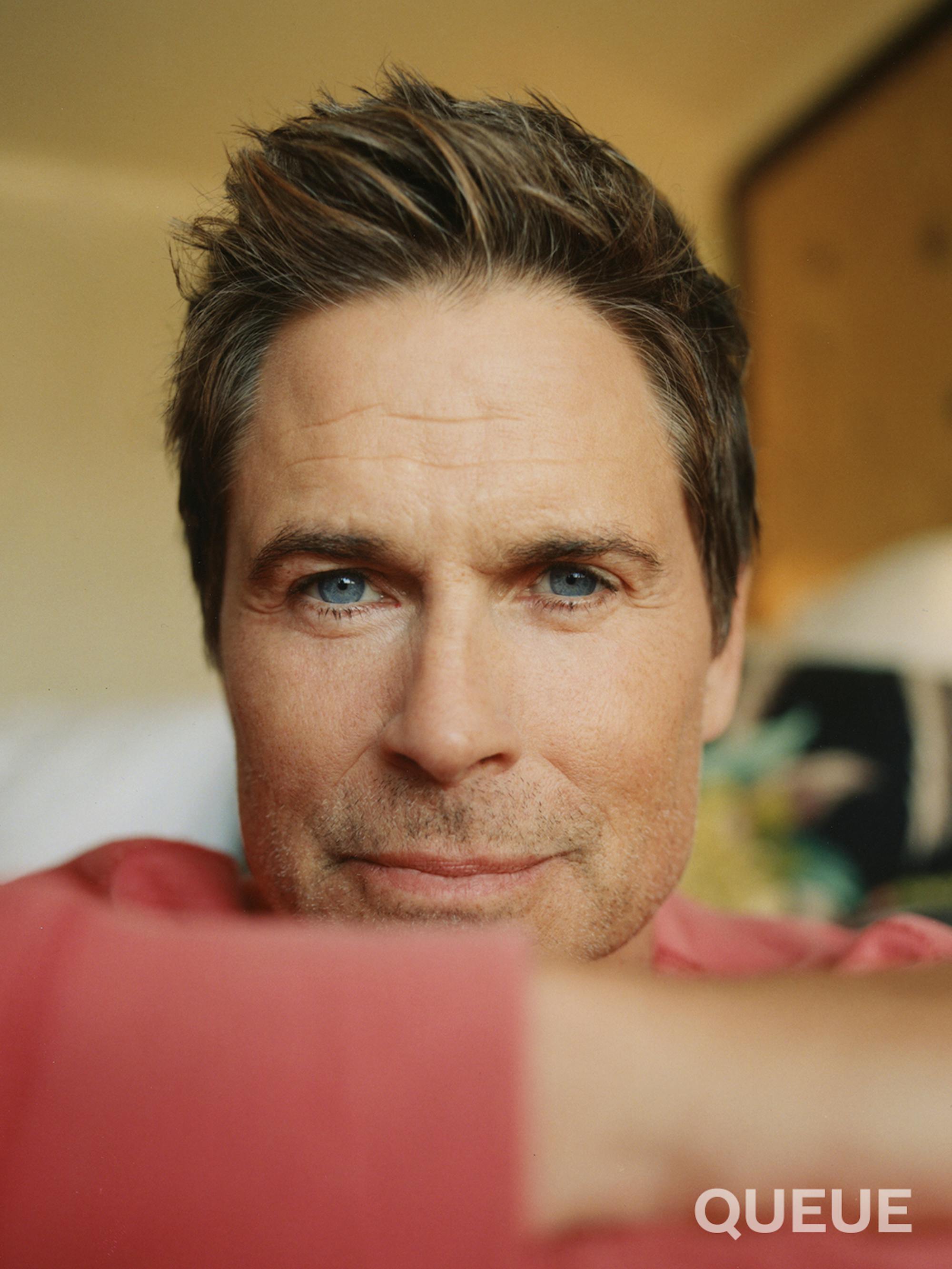 Rob Lowe gets close to the camera, smizing. He wears a red sweater.