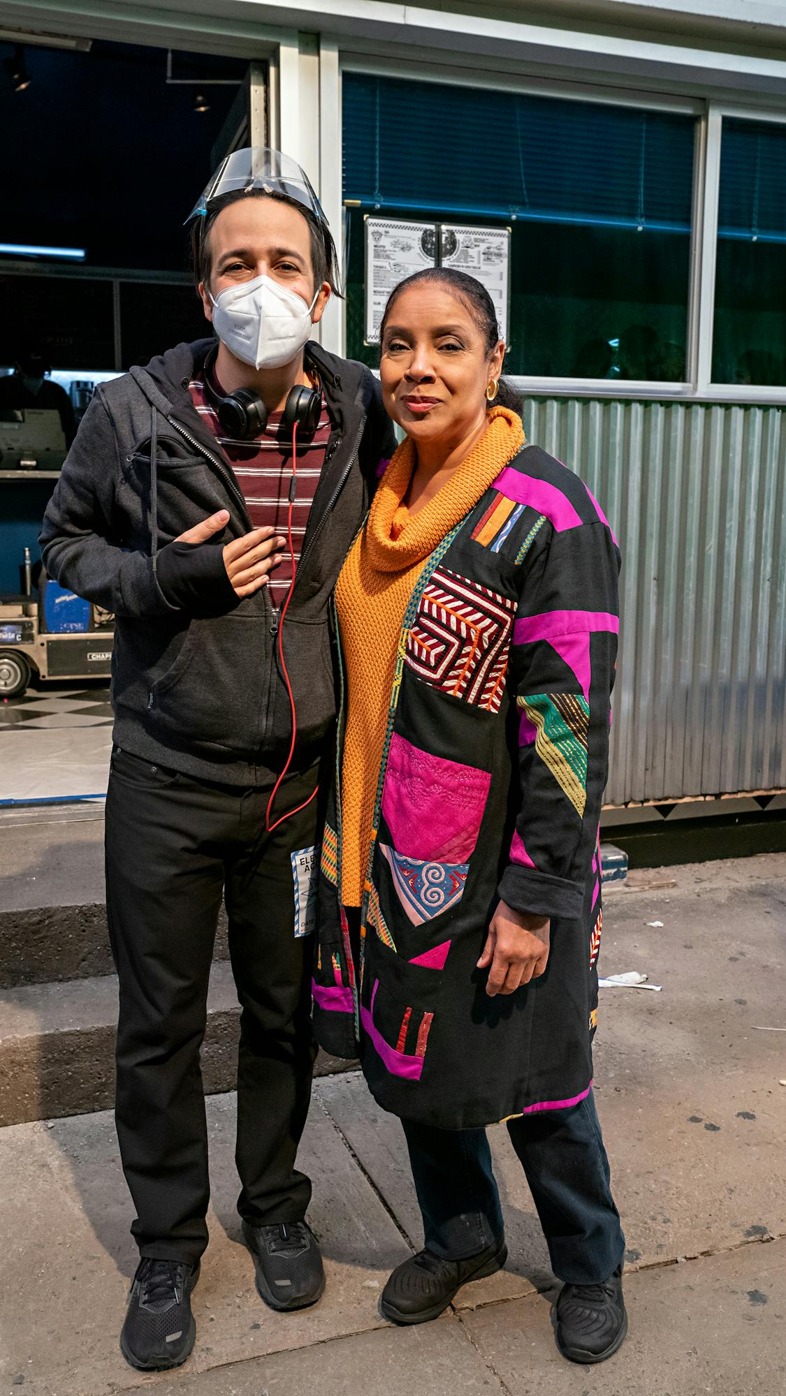 Lin-Manuel Miranda and Phylicia Rashad stand outside the Moondance Diner. Miranda wears dark clothing and protective gear. Rashad wears a colorful sweater and orange turtleneck.