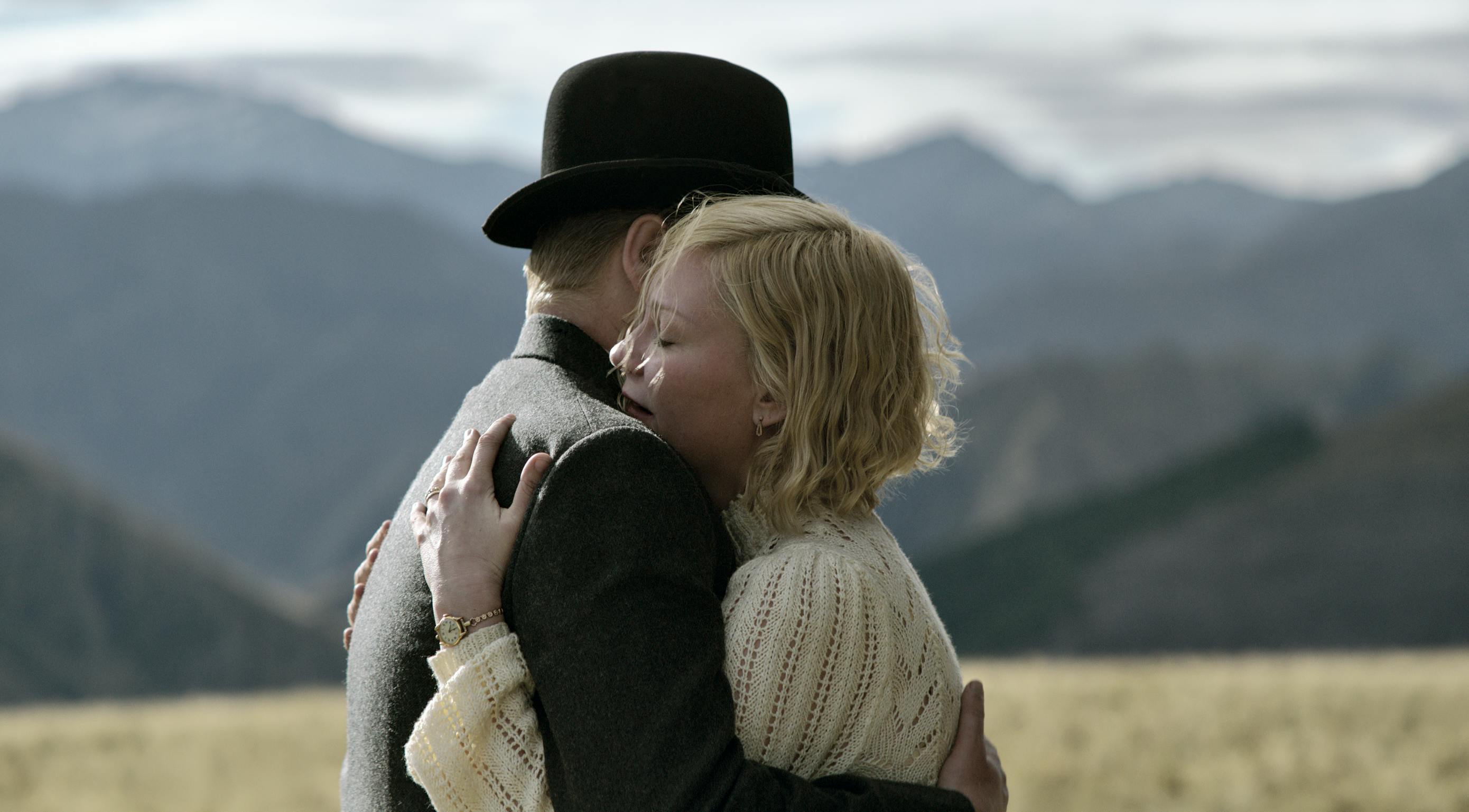 Jesse Plemons and Kirsten Dunst hug in the middle of an open field. He wears a dark suit and hat, she wears a sheer white blouse. The mountains behind them are blue-green, and blurry.