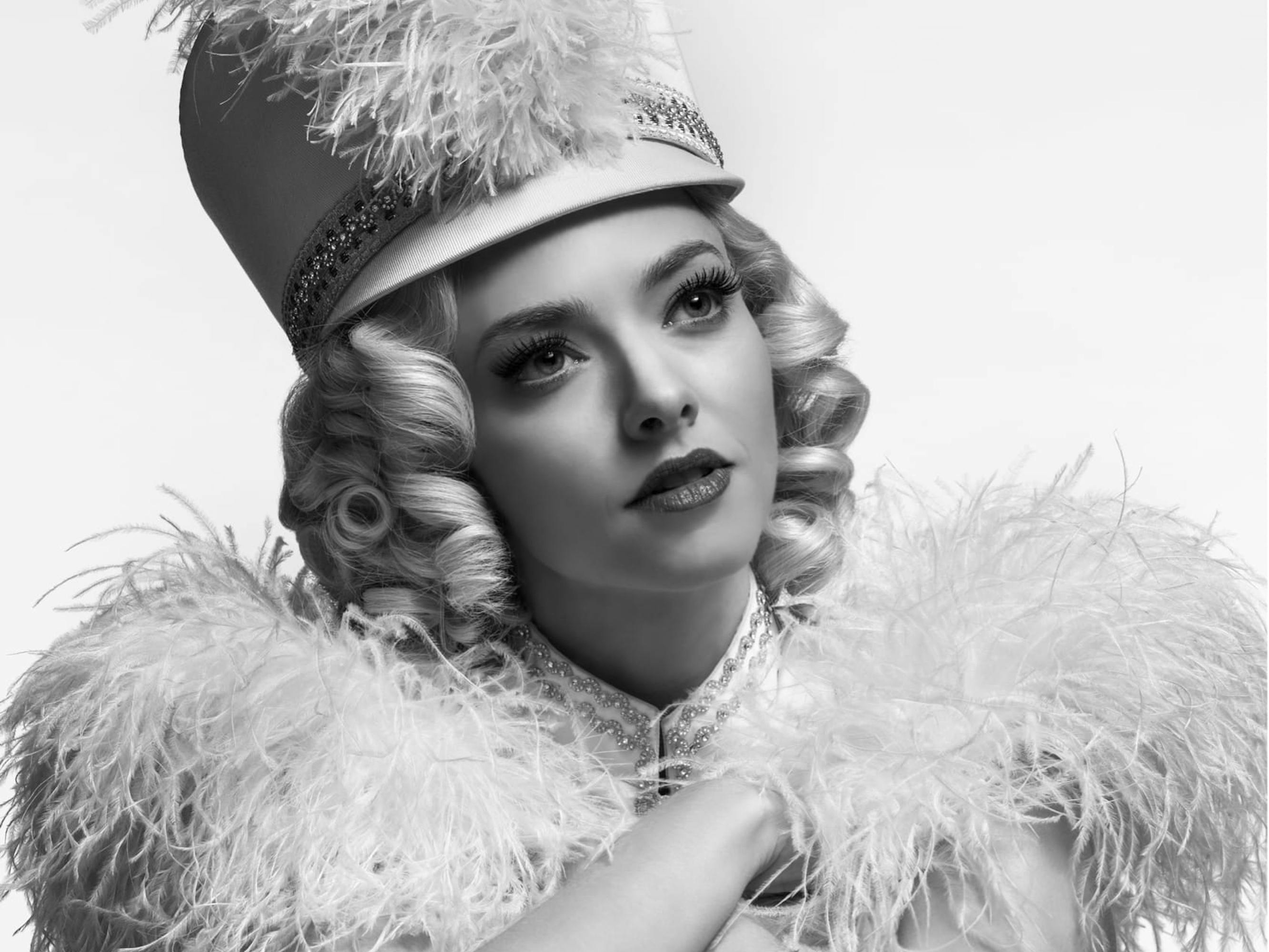 A portrait of Seyfried as Marion Davies. She’s pictured against a white background and lit in that soft Old Hollywood glow. Her blonde hair is in ringlets and she wears a cap with a prominent feather down the middle, plus matching white feathery epaulettes.